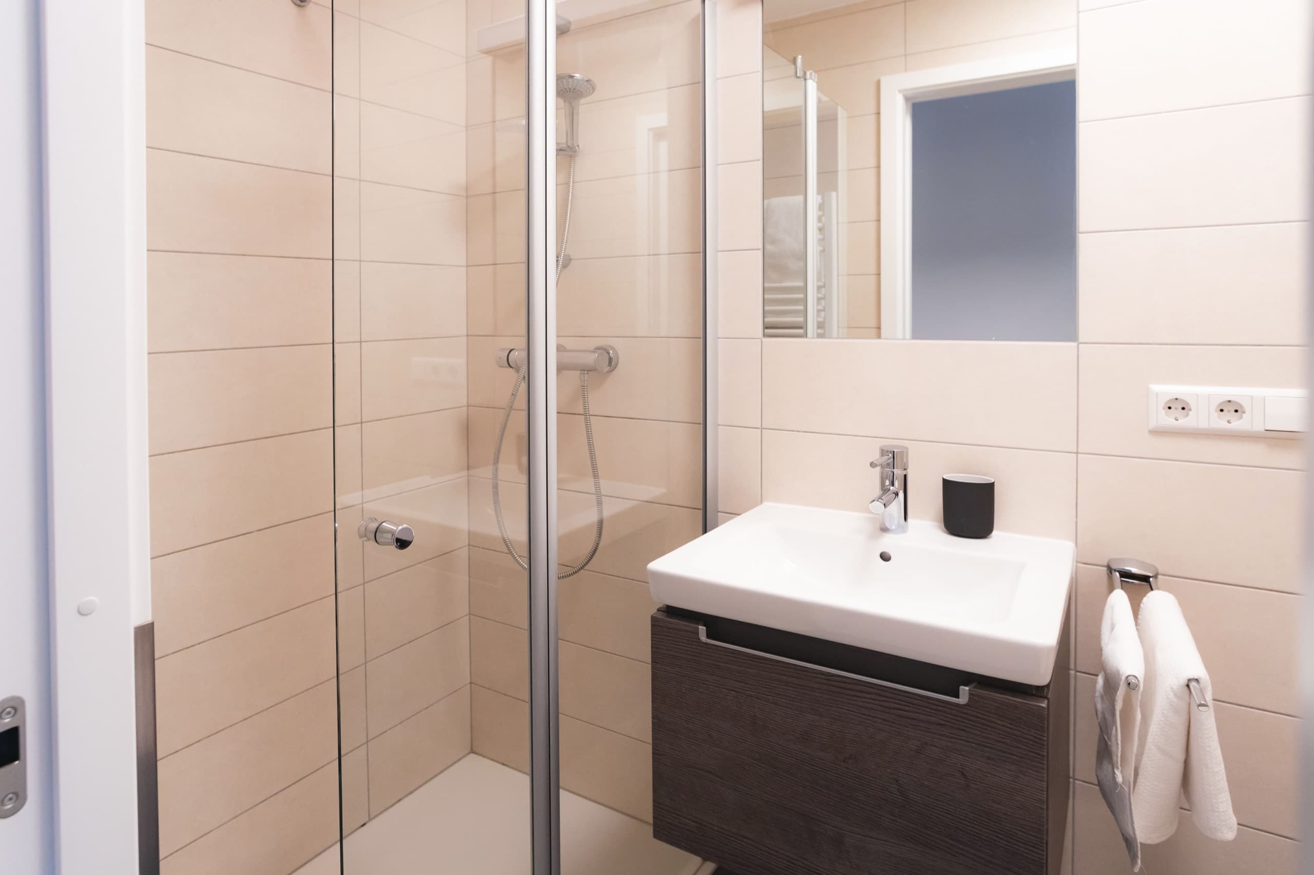 Bathroom with a shower cabin and a white sink next to it. Above the sink, there is a cabinet with mirror doors. Towels and soap are included.