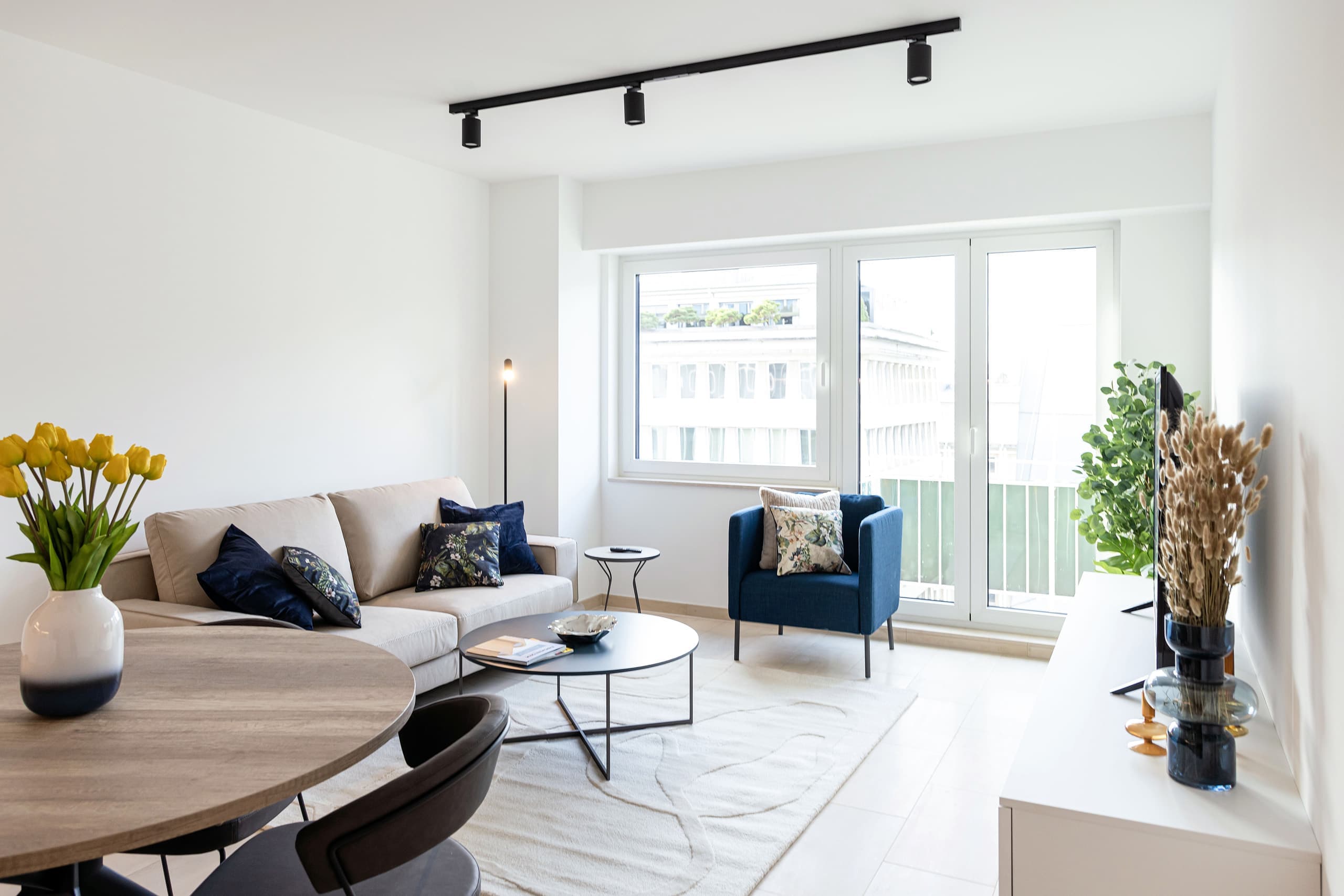 Living room area with a sofa with cushions, a coffee table with a white carpet underneath, and a standing lamp. In front of the balcony door, there is an armchair with cushions on it.
In front of the sofa, there is a TV on a TV stand.