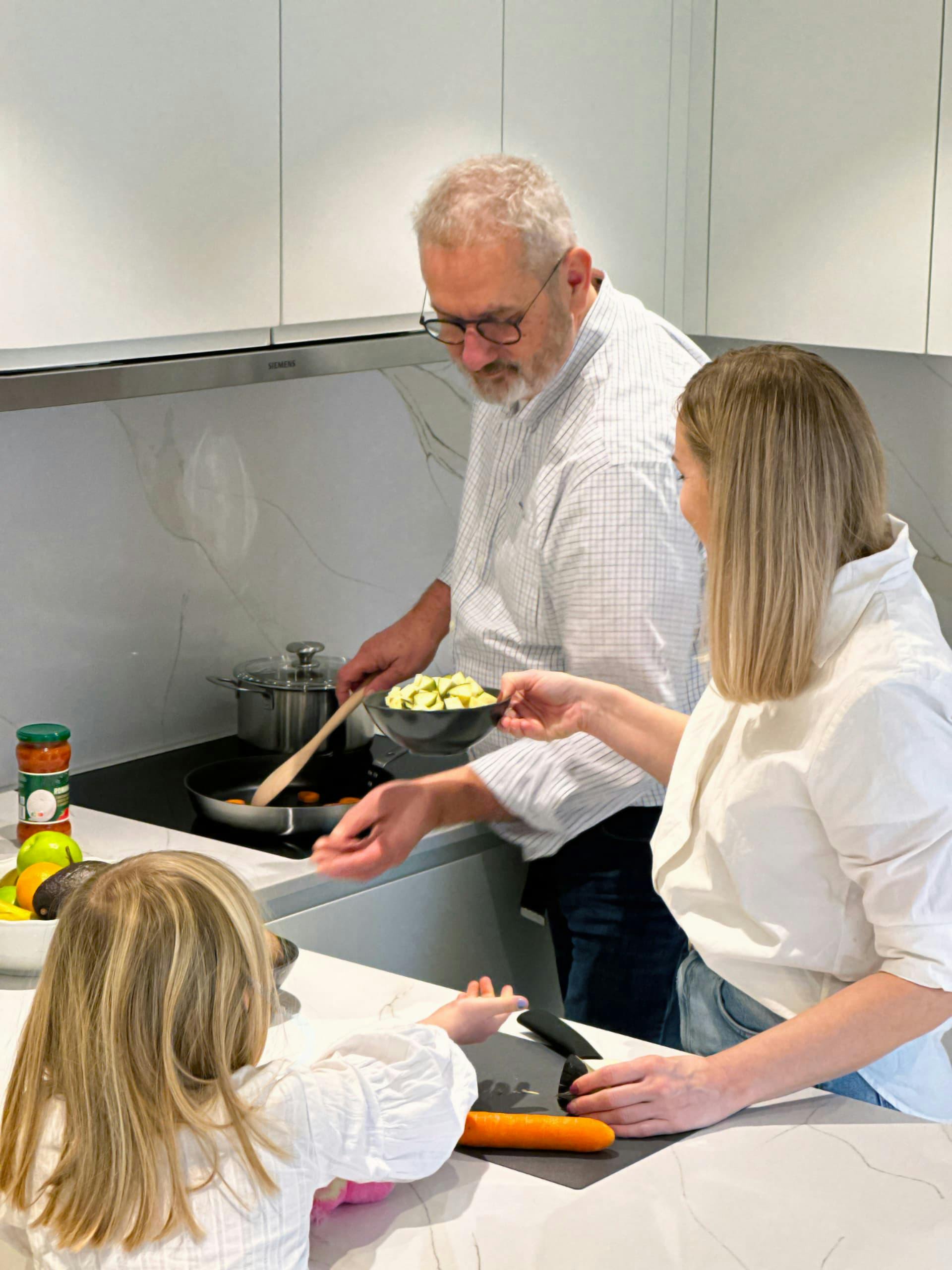 The picture depicts a family cooking in a kitchen. It shows the electric stove, a cooking pan on top of it with vegetables inside, and on the countertop ingredients for cooking.