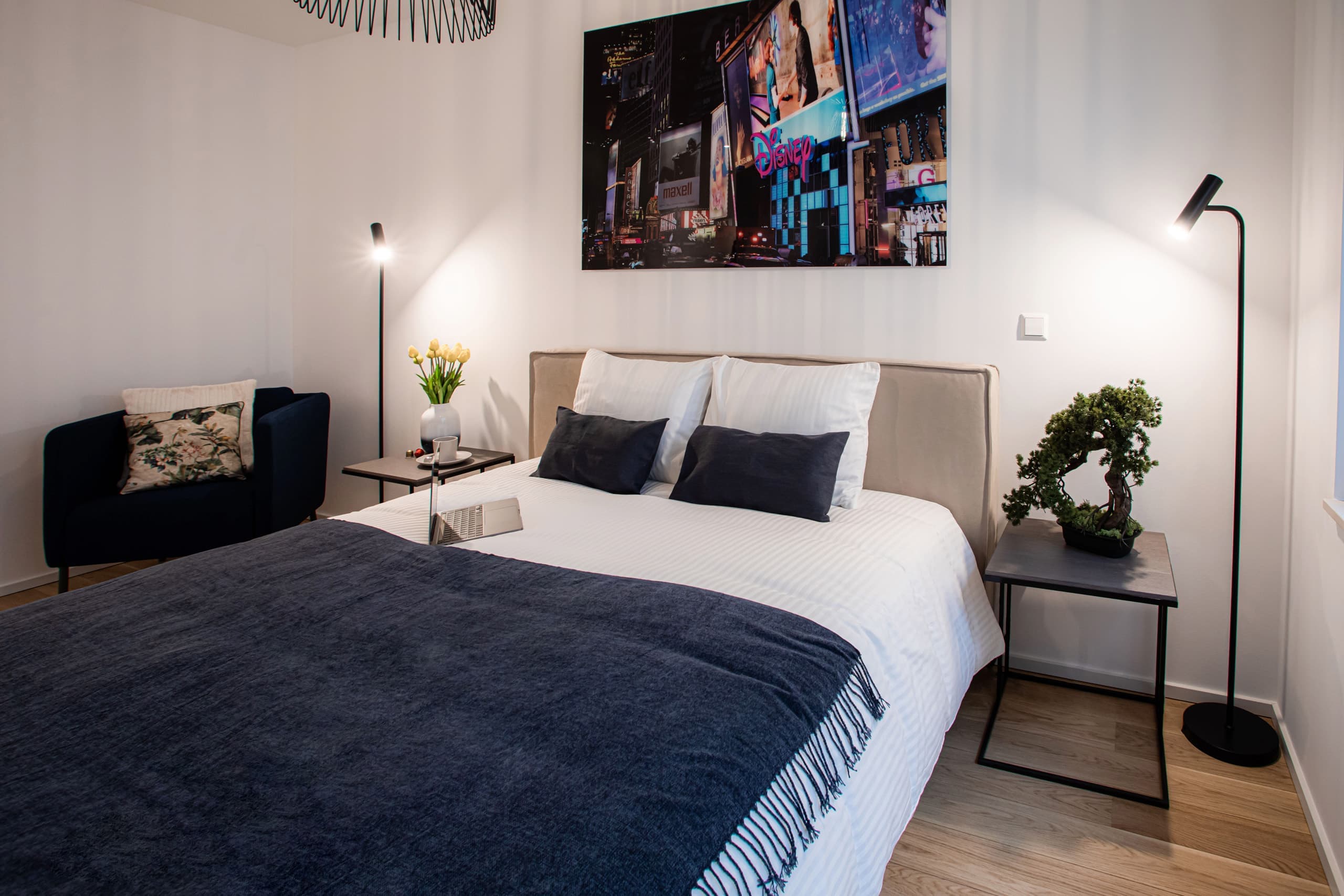 Bedroom with a large bed with white sheets. On the bed, there is a laptop. Two lamps and two nightstands flank the bed. On its left, there is an armchair.