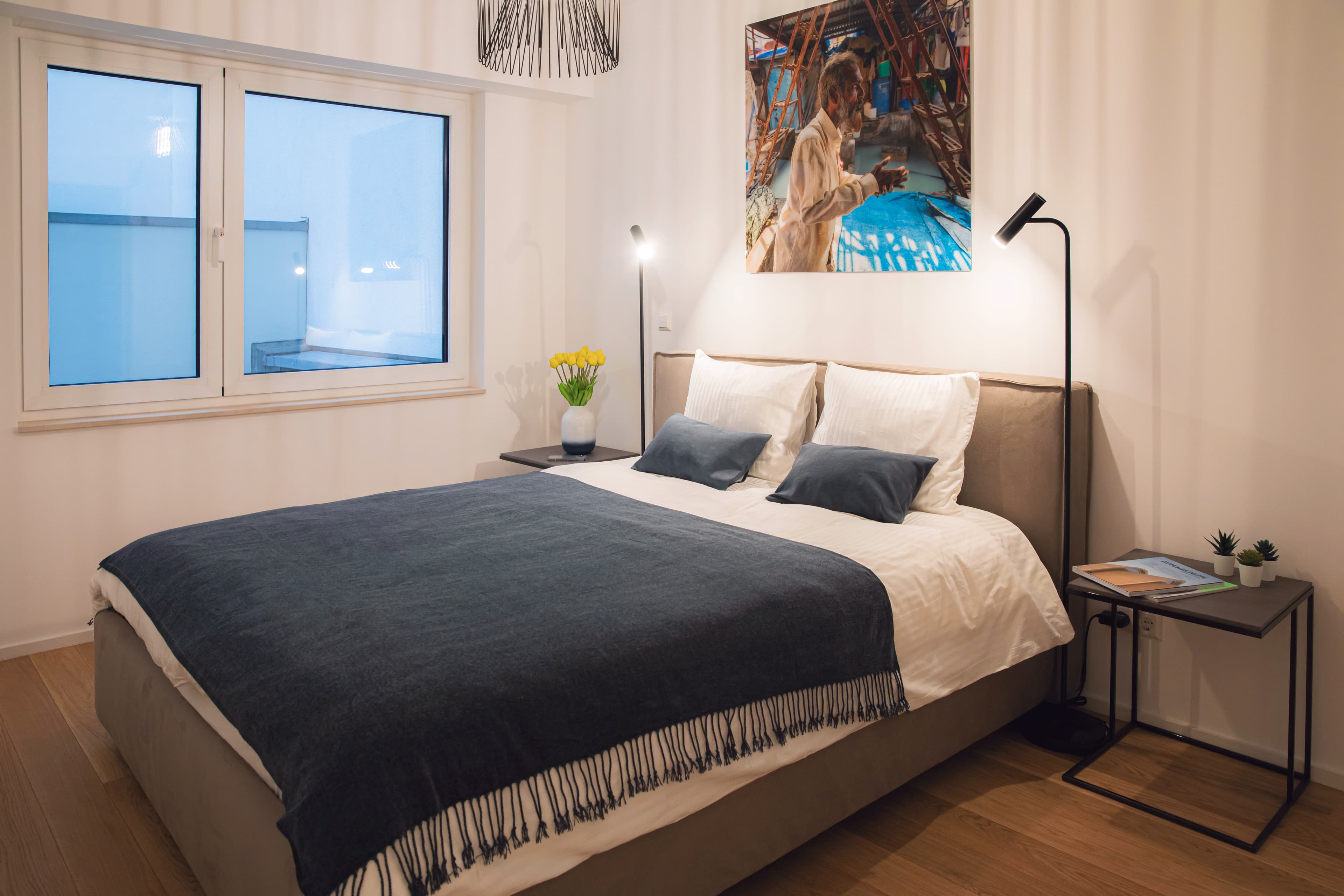 Bedroom with a large bed flanked by two lamps and two nightstands. On the right, there is a large window, and above the bed hangs a big picture.