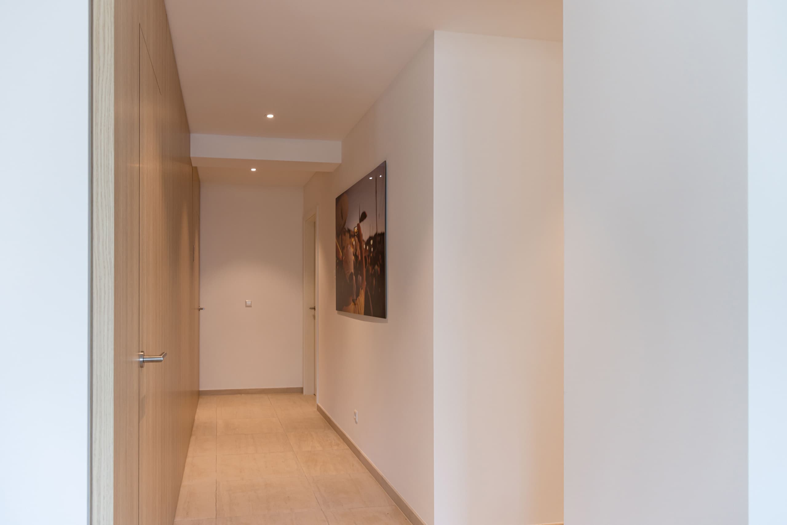 Long hallway with picture hanging on the wall, and two doors on the left. The wall is covered in light wood.