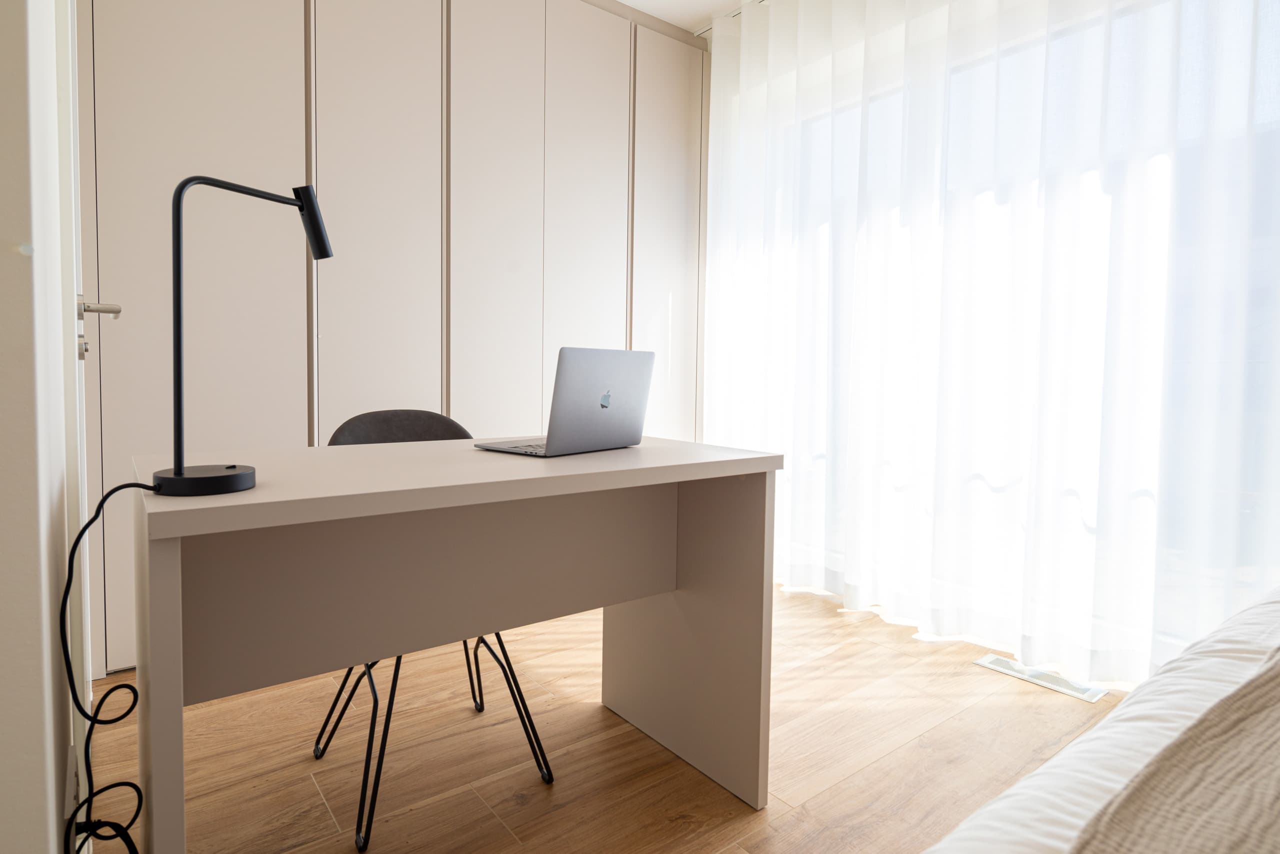 Desk with laptop and lamp, natural light through a ceiling-to-floor window on the right. Behind, a large wardrobe and a chair.
