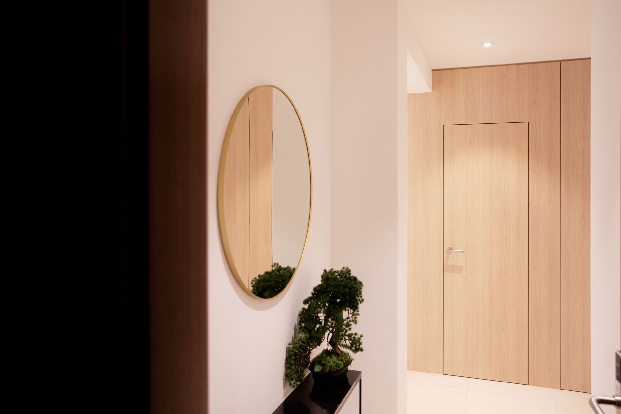 Hallway view, with entry door in the back. On the wall, a wooden frame oval mirror with a small bonsai tree underneath.