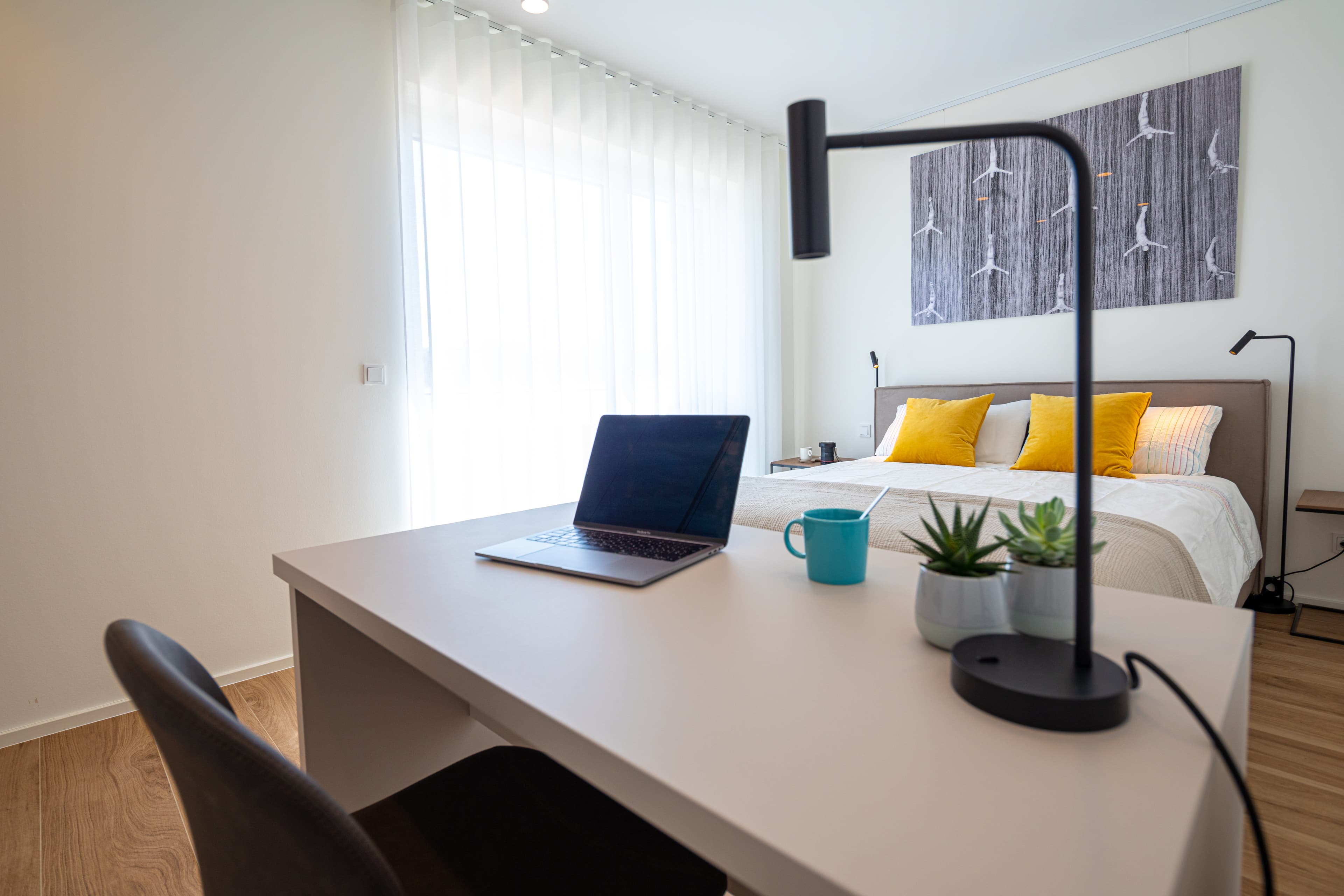 Bedroom with a large bed with white sheets and mustard cushions, flanked by two lamps and nightstands. In front is a desk and chair, with a laptop, coffee cup, and two small flower pots.