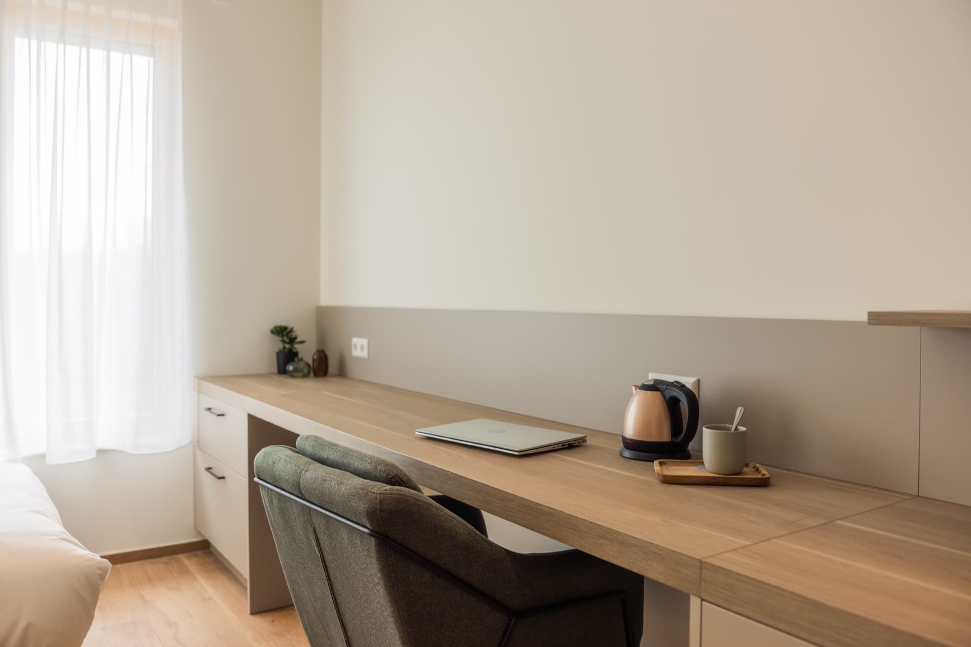 The picture depicts a close-up image of the working area, with a large desk with storage drawers next to it. On top of the desk there is a kettle and a cup and a small vase with a plant in it