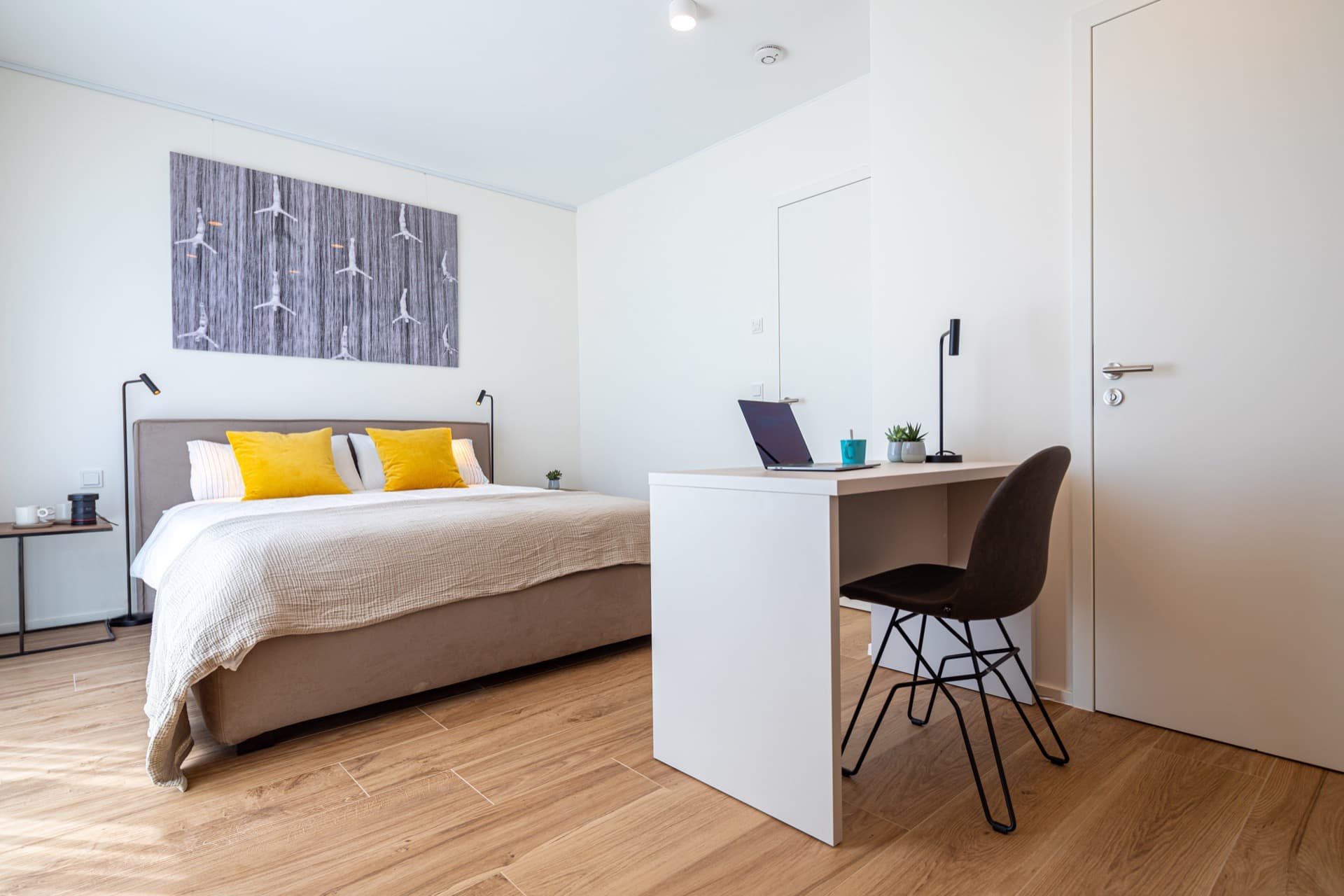 Bedroom with a king-size bed with white sheets and mustard cushions, flanked by two nightstands and two lamps. In front, a desk with a chair. On the desk, there is a laptop, a lamp, a coffee cup, and two small flower pots with aloe vera. On the right side of the desk is a door.