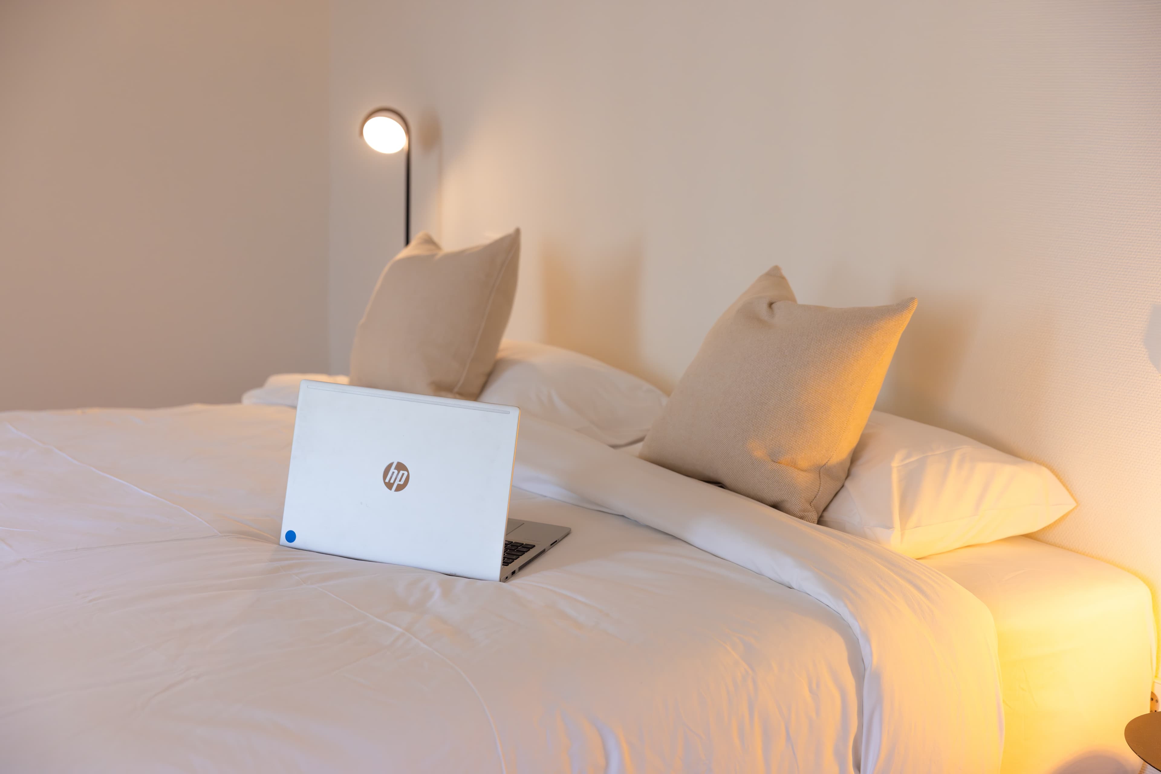The picture depicts a close-up of the bed with cotton white linens and cushions. On top of the bed there is an open HP Laptop