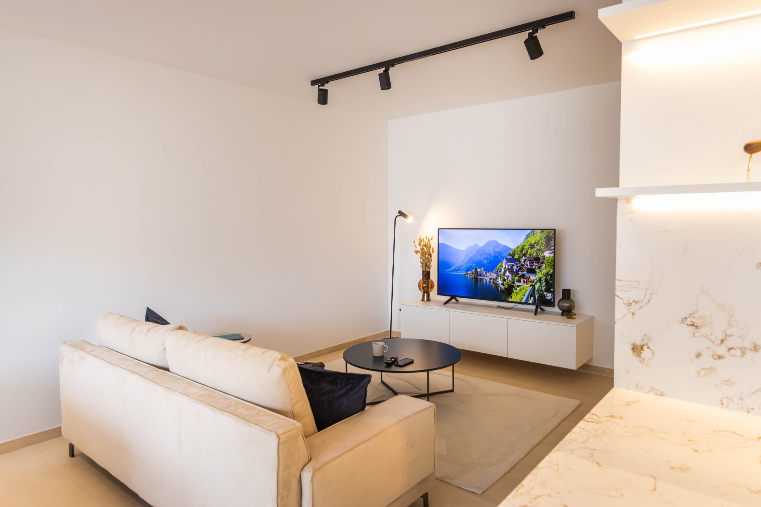 View from the kitchen of the sofa's back, adorned with a coffee table and a white rug beneath. In front, there's a TV stand supporting a smart TV, and next to it is a standing lamp.