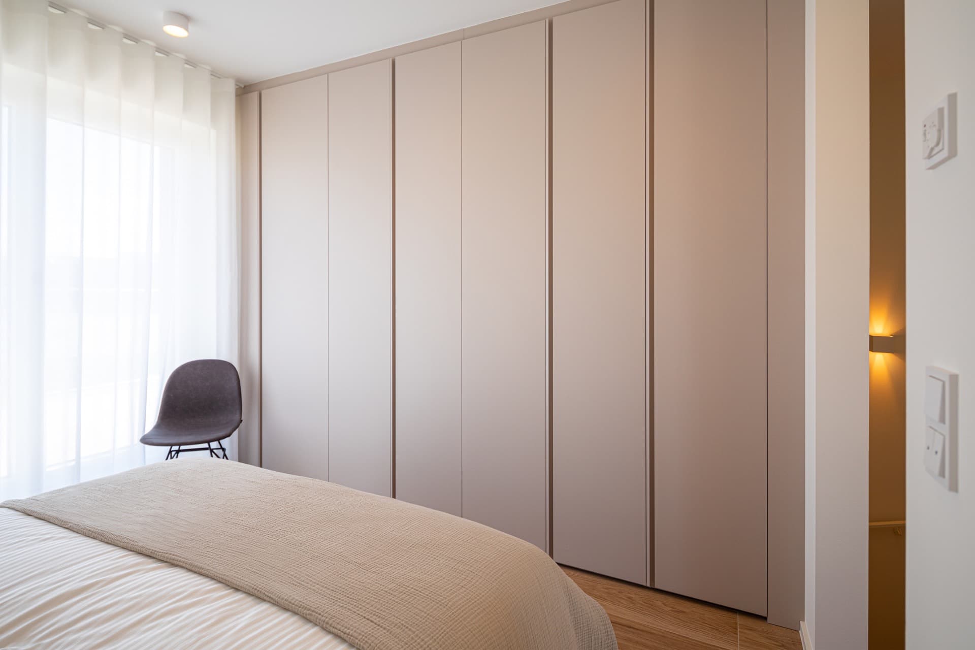 Bedroom with a wall-to-wall wardrobe in front of a large bed. There is a chair in the corner next to the large window.