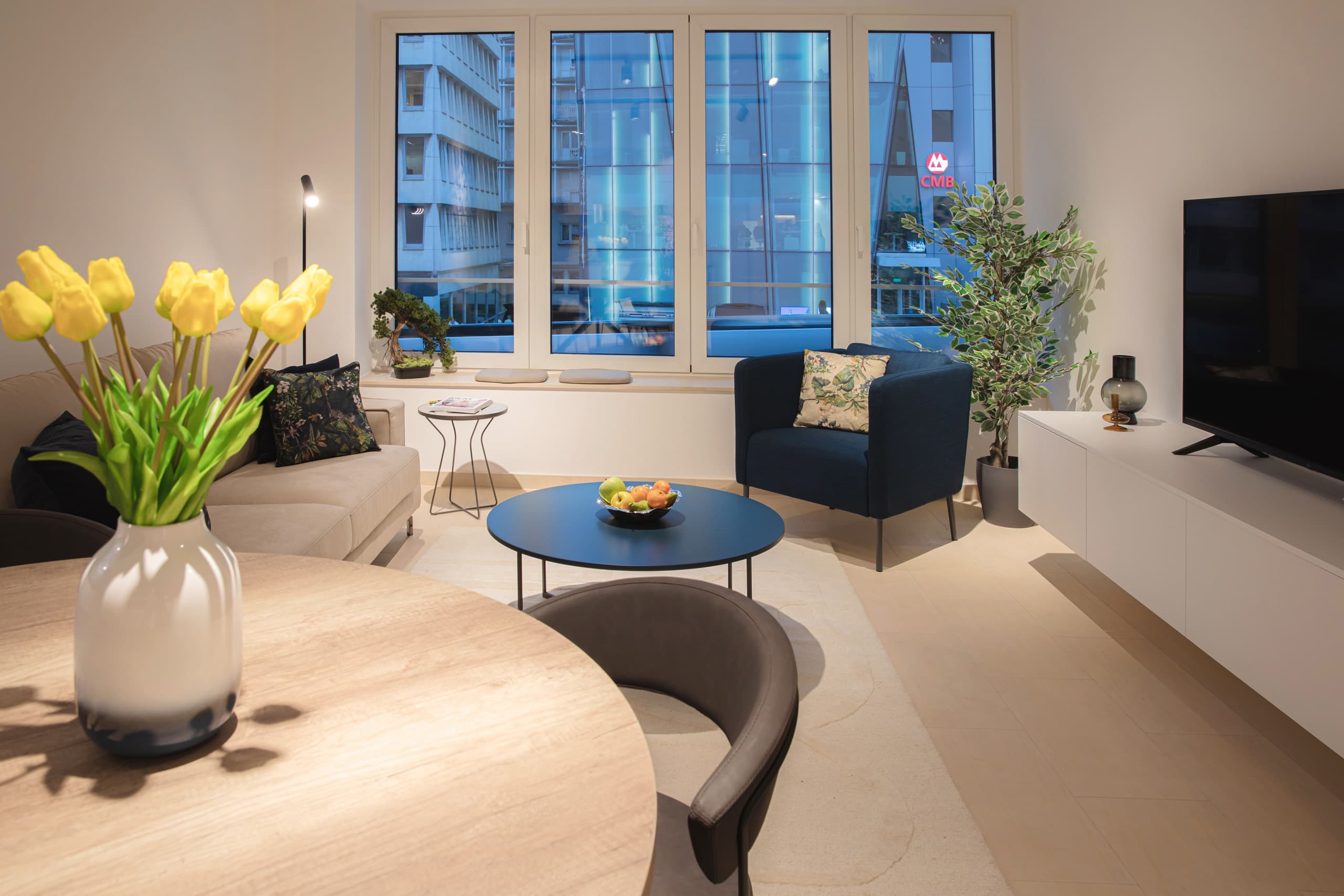 The image shows a circular living room table decorated with a vase of yellow tulips and three chairs. To its left is a couch with cushions, and in front of it, a small coffee table with a fruit bowl. Positioned in front of the small table is a dark blue armchair. Adjacent to it is a white TV stand with a TV.