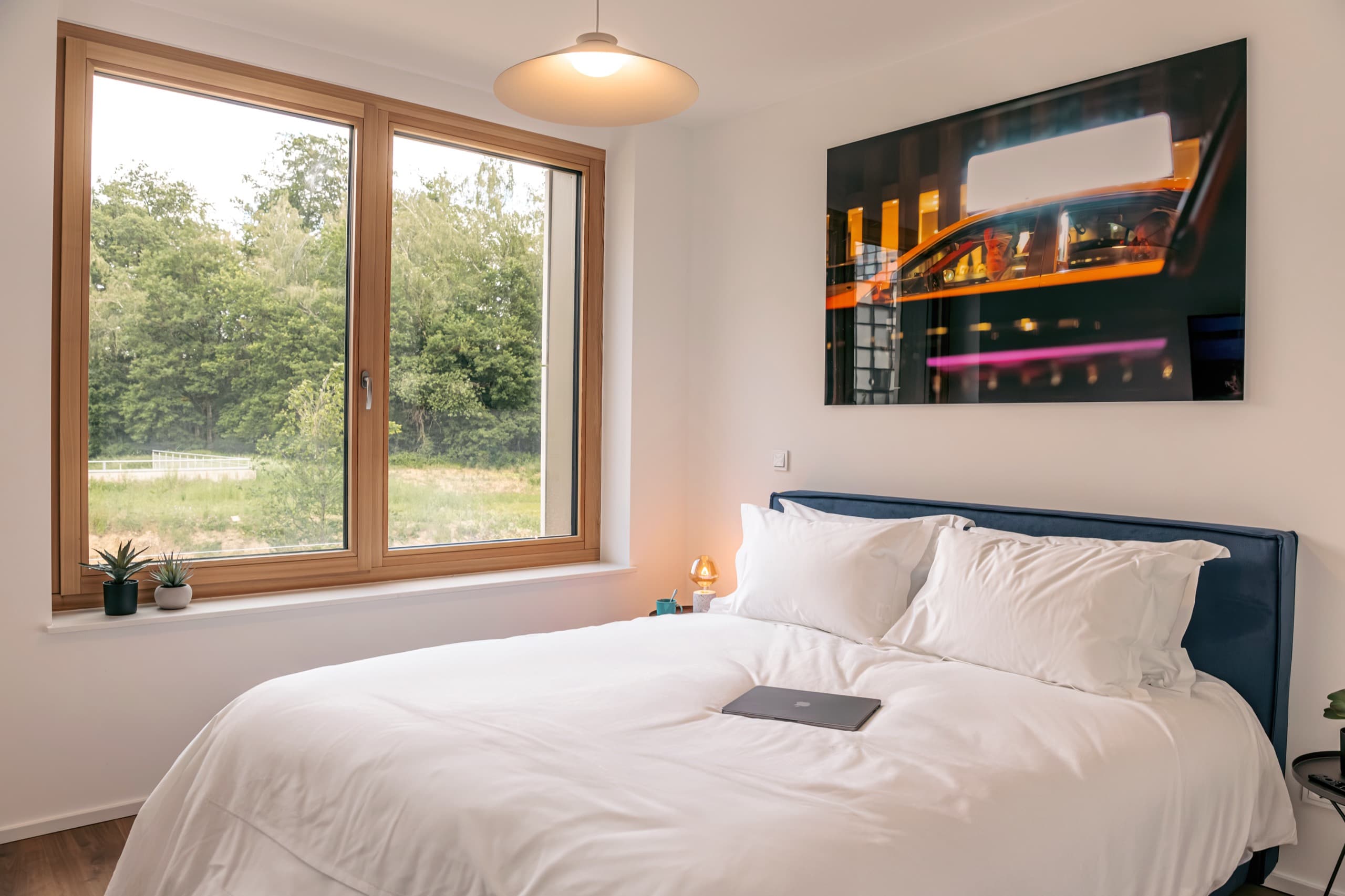 Bedroom on the ground floor, with a large window overlooking the forest. King-size framed bed with cotton white sheets. A framed big picture hangs above the bed. Night stand and table lamp with warm light are next to the bed.