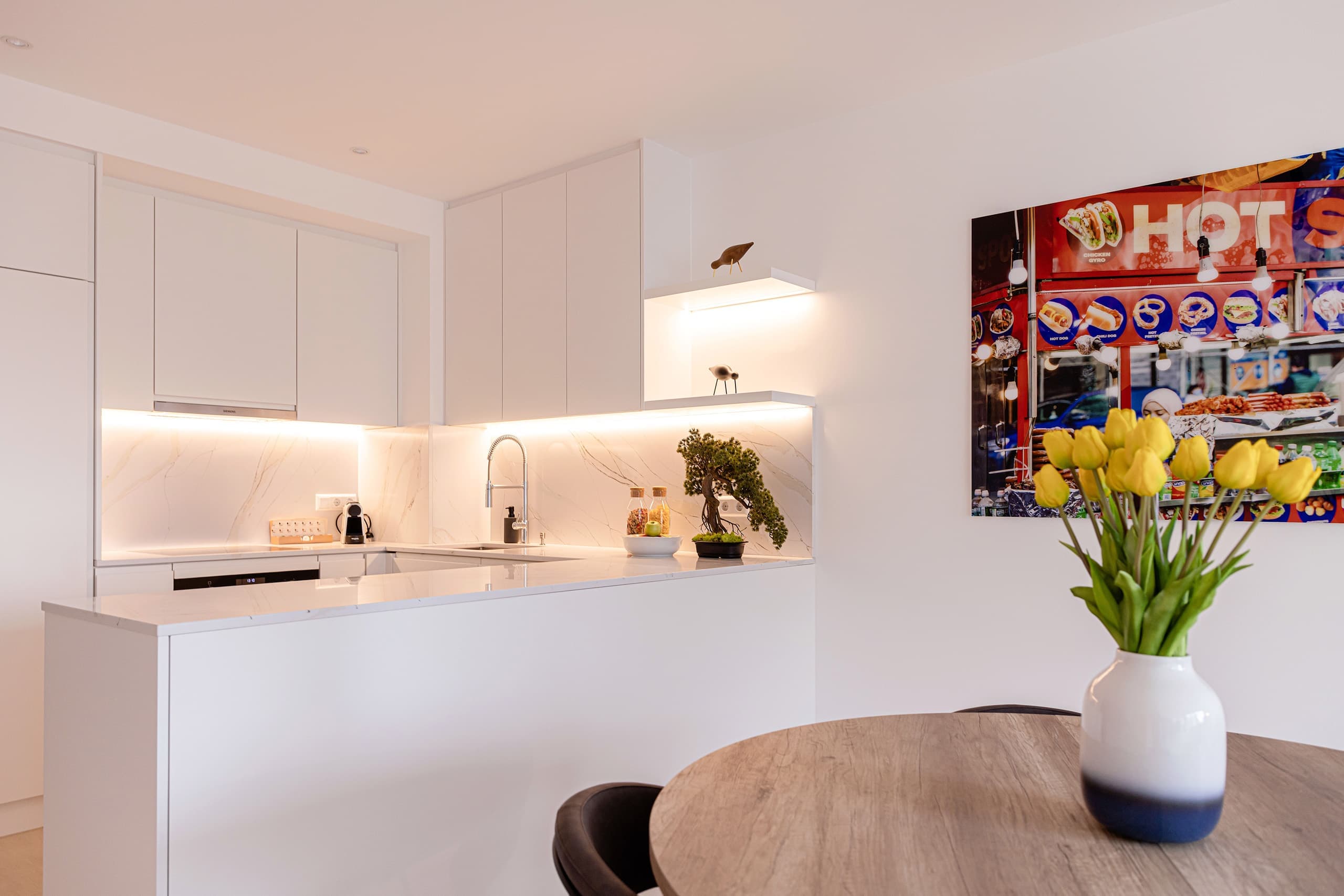 The kitchen area, fully equipped. In front of it, there is a dining table with chairs and a flower vase on it. On the wall, hangs a big picture