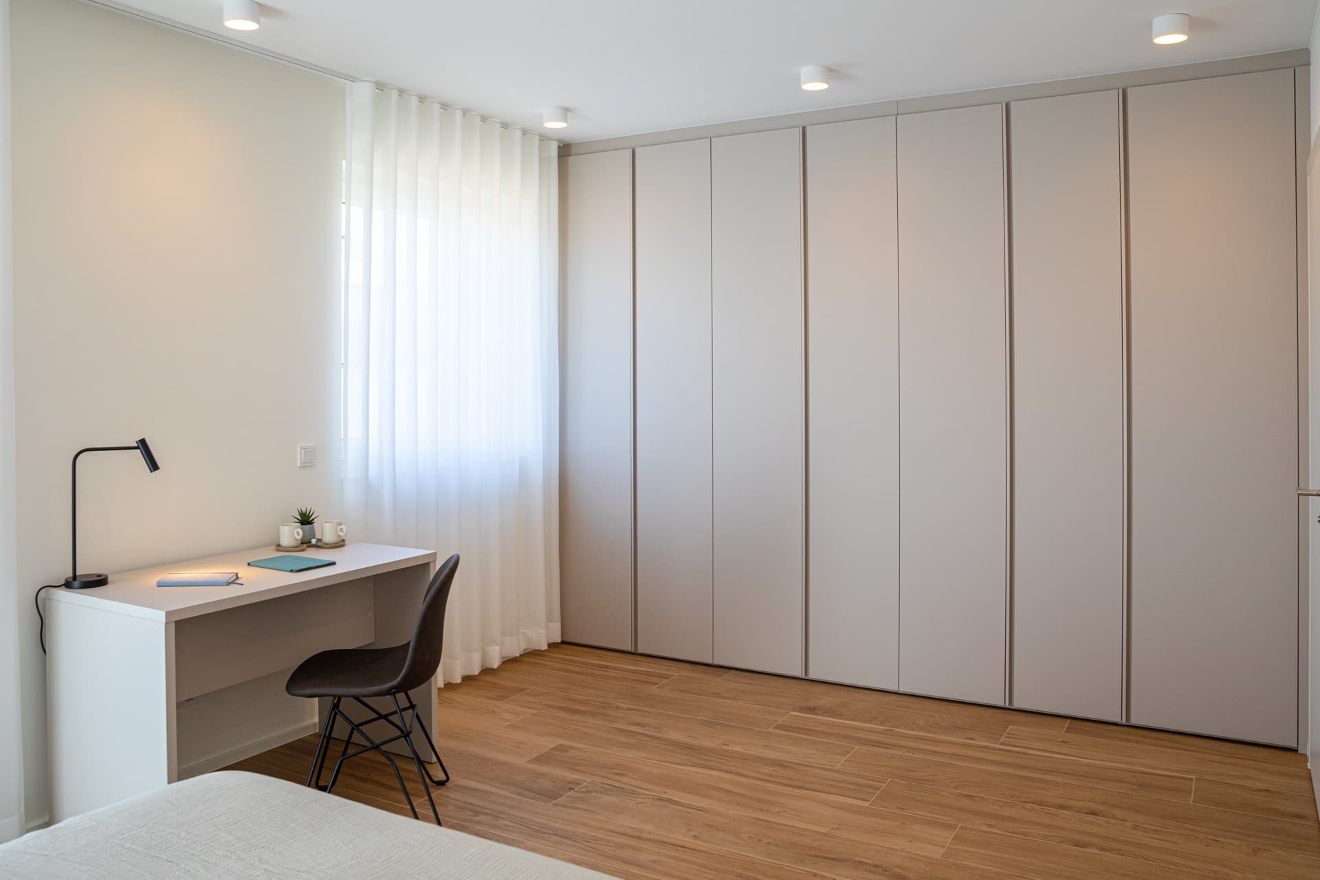 Picture of the room corner with a large wardrobe, a large window with white curtains, and a desk with a chair on the left. On the desk, there's a tablet, an agenda, two coffee cups, and an aloe vera plant.