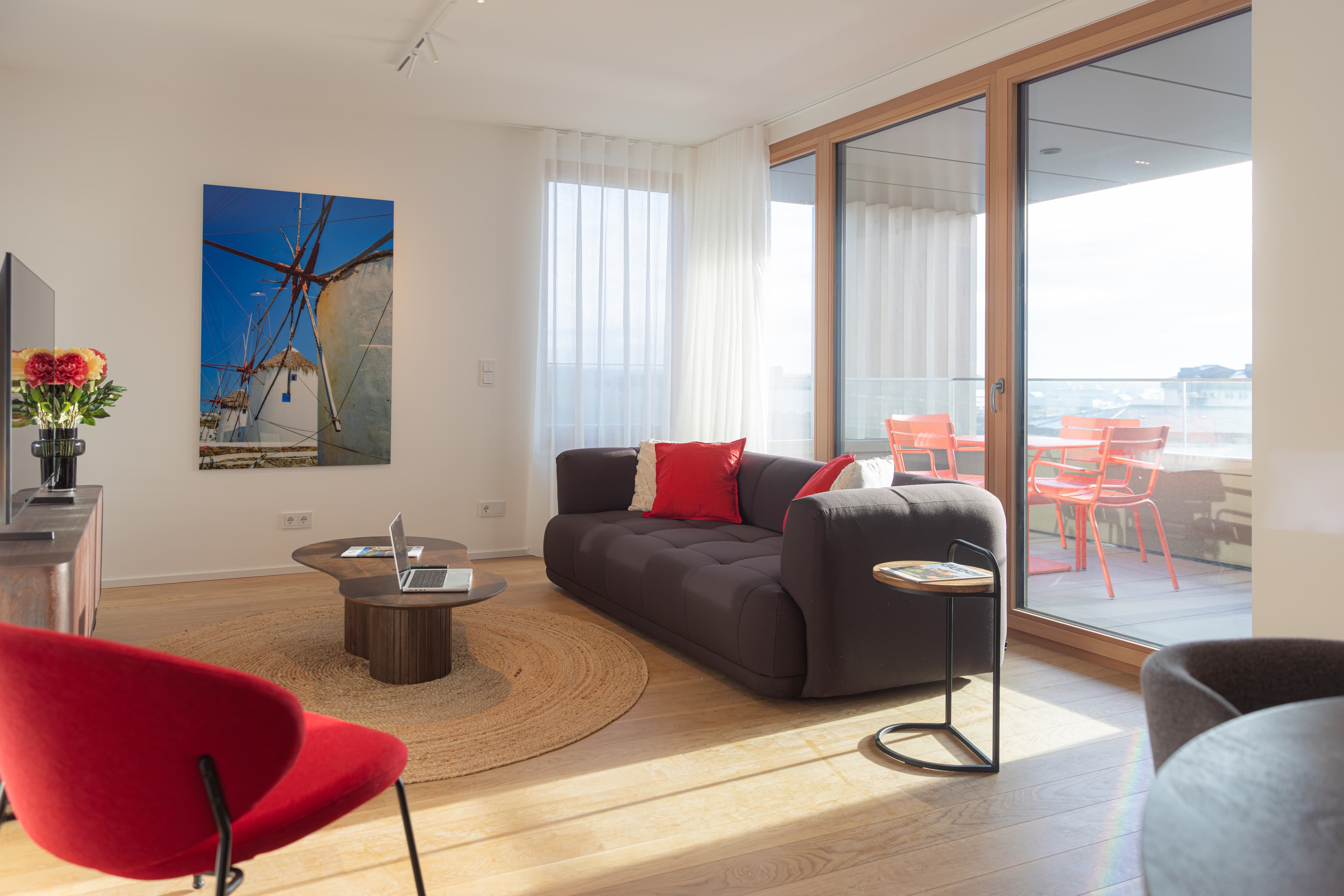 Overview of the lounge area with a large comfortable sofa, plush cushions, and a small table next to it.
In front, there is a coffee table with a laptop on top. In front, there is a TV stand with a large LCD screen on top.
Behind the sofa, there is the large balcony, with floor-to-ceiling windows, a red outside table, and chairs.