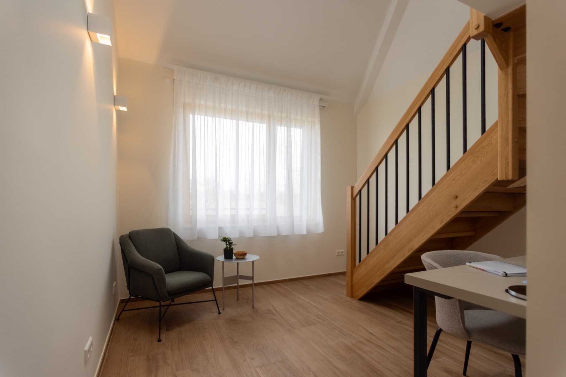 The picture shows the downstairs area of a furnished bedroom for rent in Luxembourg city. It contains a working area with a desk and a comfortable chair, an armchair with a coffee table next to it, and, behind, there is a large window flooding the room with natural light.
