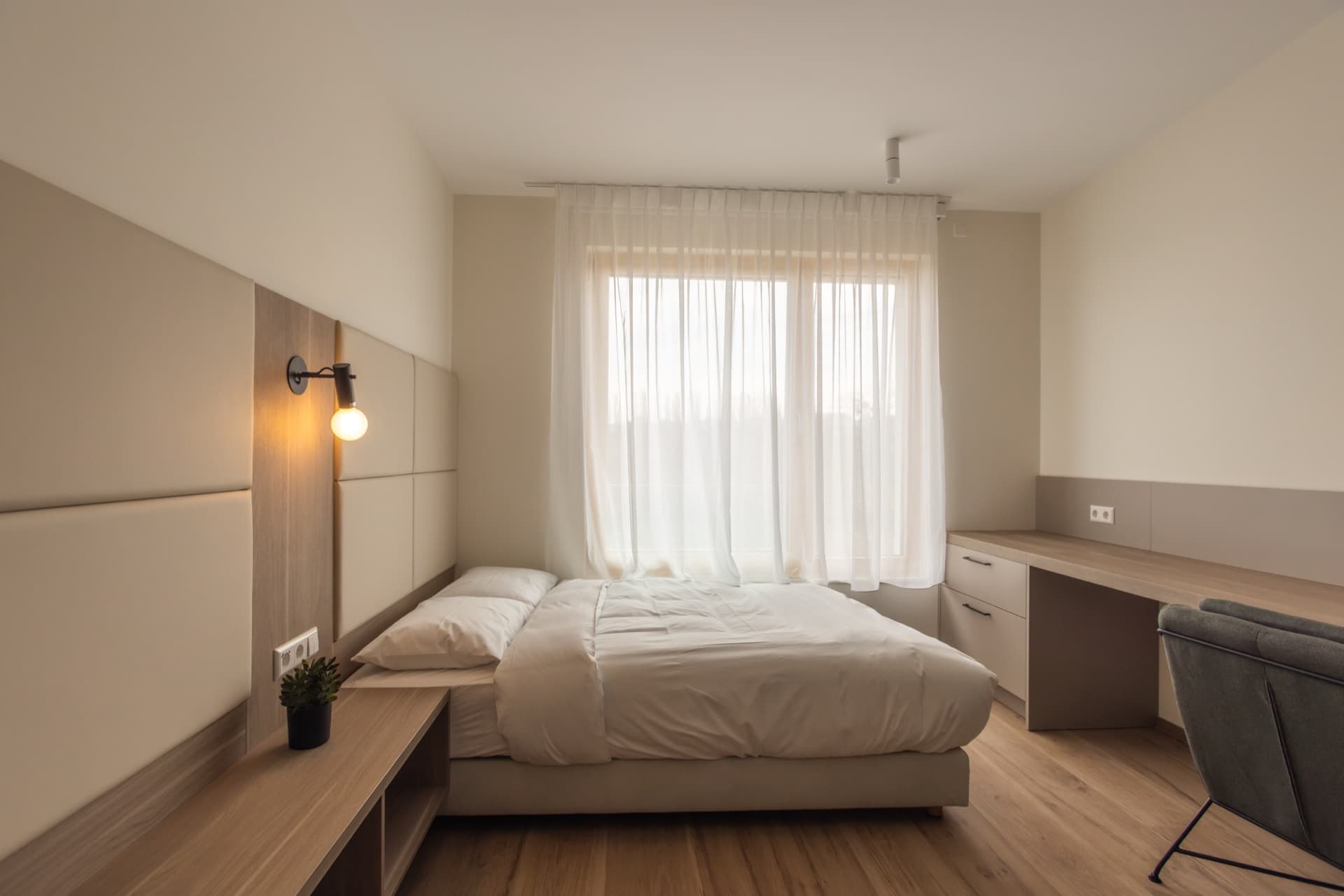 Overview of the bedroom with a framed king-size bed, a desk and a rollbox with drawers, and large windows flooding the room with natural light.