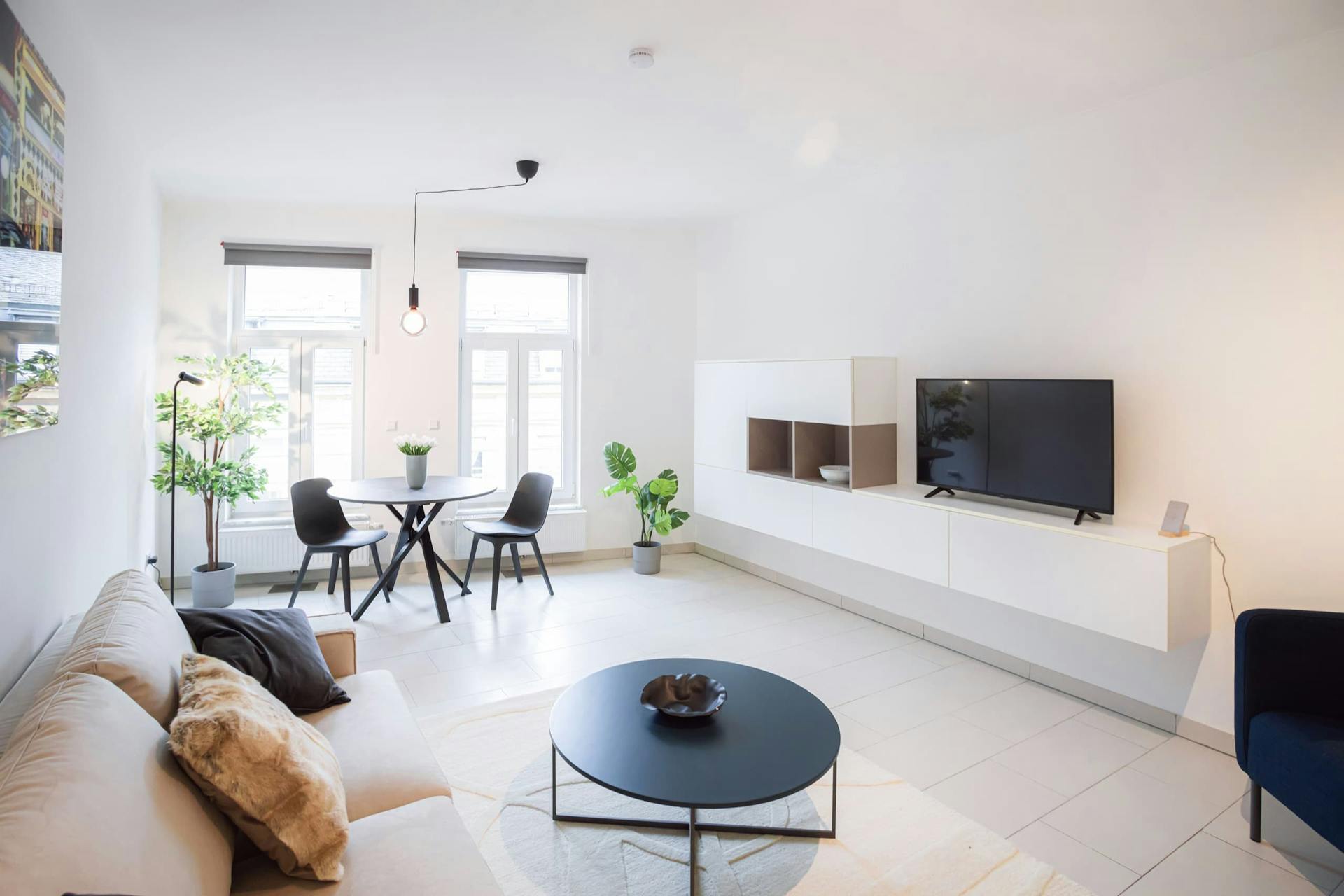 Lounge area comprised of a sofa with cushions and a coffee table with a white carpet underneath. In front of the sofa is a TV stand with a smart TV on it. In front of two floor-to-ceiling windows is a dining table and chairs.