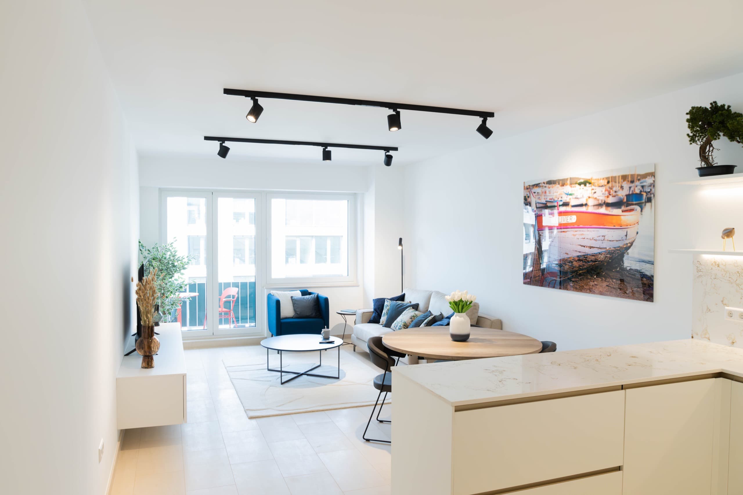 Lounge area view from the kitchen. On the left, a TV stand with a smart TV on it. On the right, a dining table with chairs and a flower vase on top. Next to it, there is a couch with cushions on it. In front of the couch, there is a coffee table with a white carpet underneath.
In front of the windows and balcony door, there is an armchair with a small coffee table next.