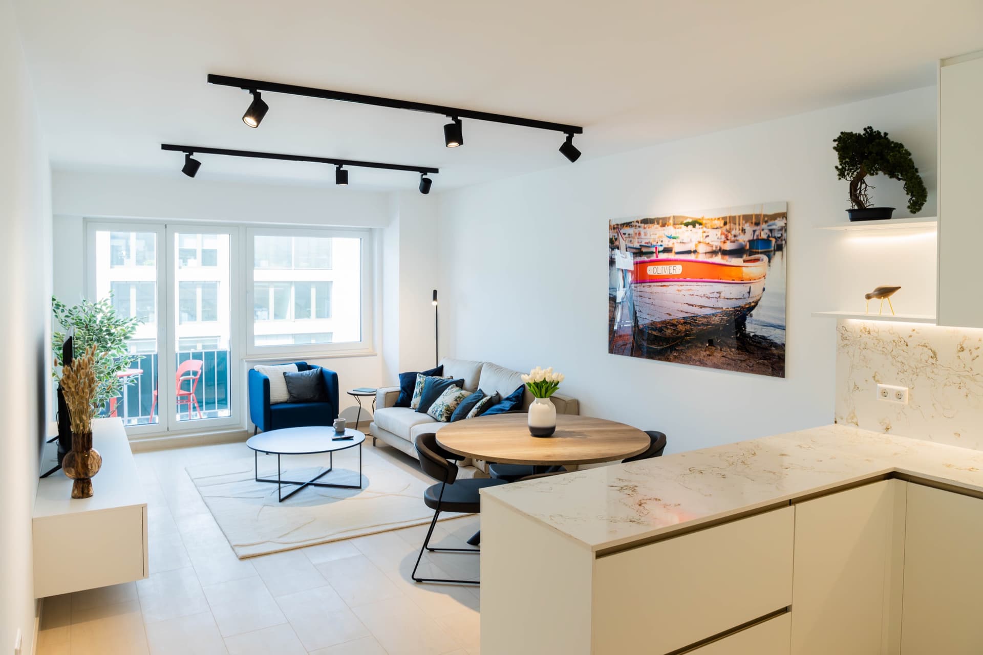 Dining area view, in front of the kitchen area. In the foreground, there is a dining table with chairs and a flower vase on top. Behind, is a sofa with cushions on it, and an armchair next. Large windows and balcony door light up the room. On the wall hangs a big picture.