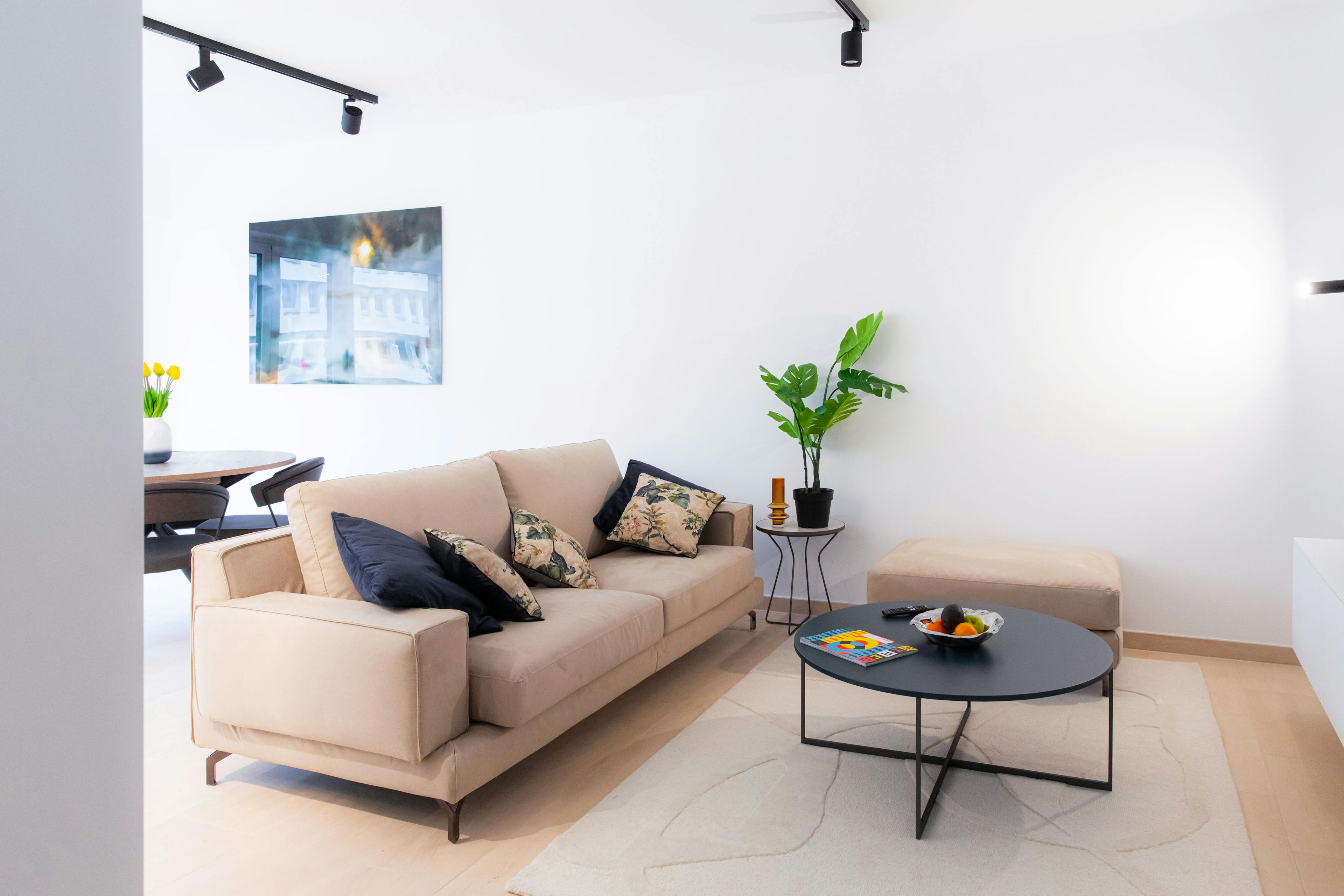 Sofa with cushions on it. In front, there's a coffee table. Behind the couch, is a dining table with chairs, next to big windows. On the ceiling hang industrial-style black lamps, and on the wall hangs a big picture. Next to the sofa, there's a small flower in a pot.