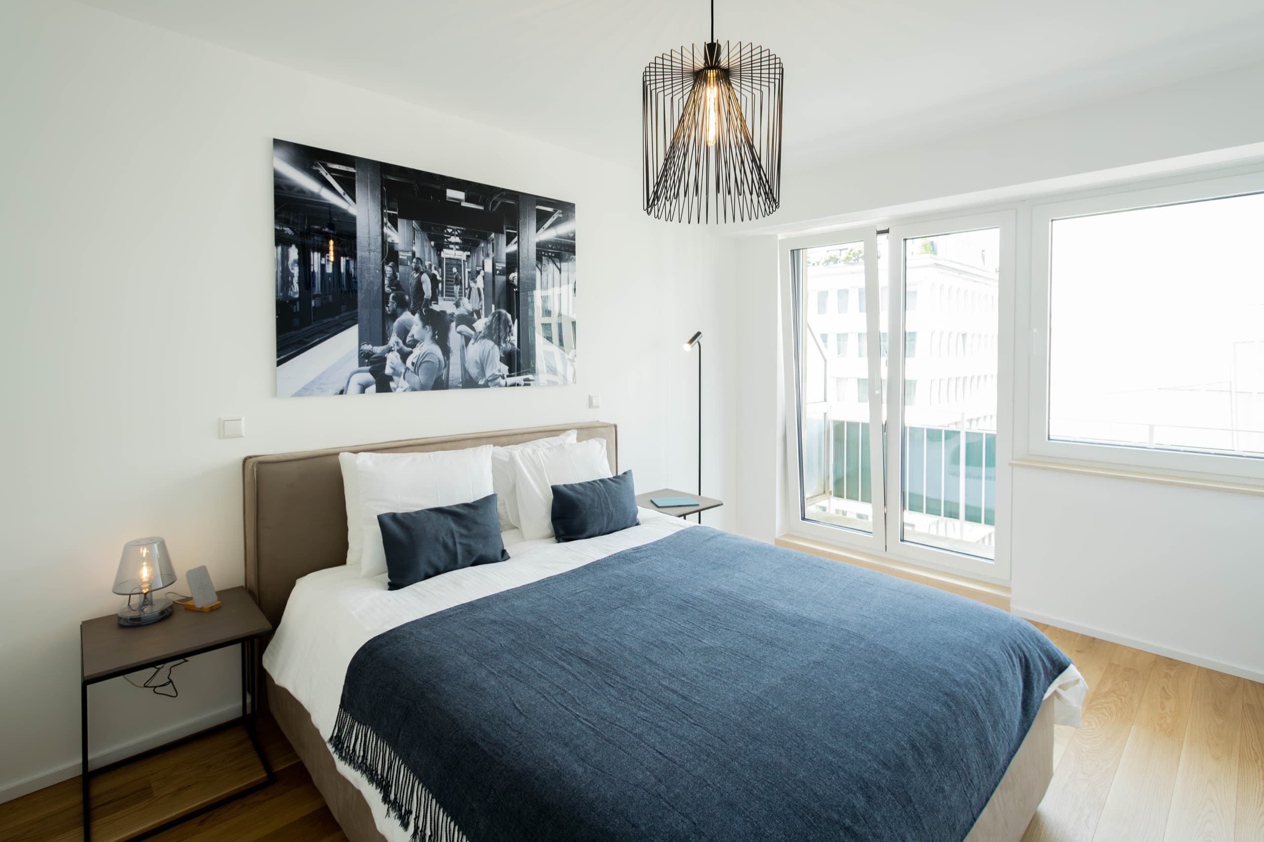 Bedroom with a large bed with white sheets and pillows. A dark blue cover on top. The bed is flanked by two nightstands and a standing lamp. Behind, on the wall, hangs a big picture. On its left, there is a floor-to-ceiling window.