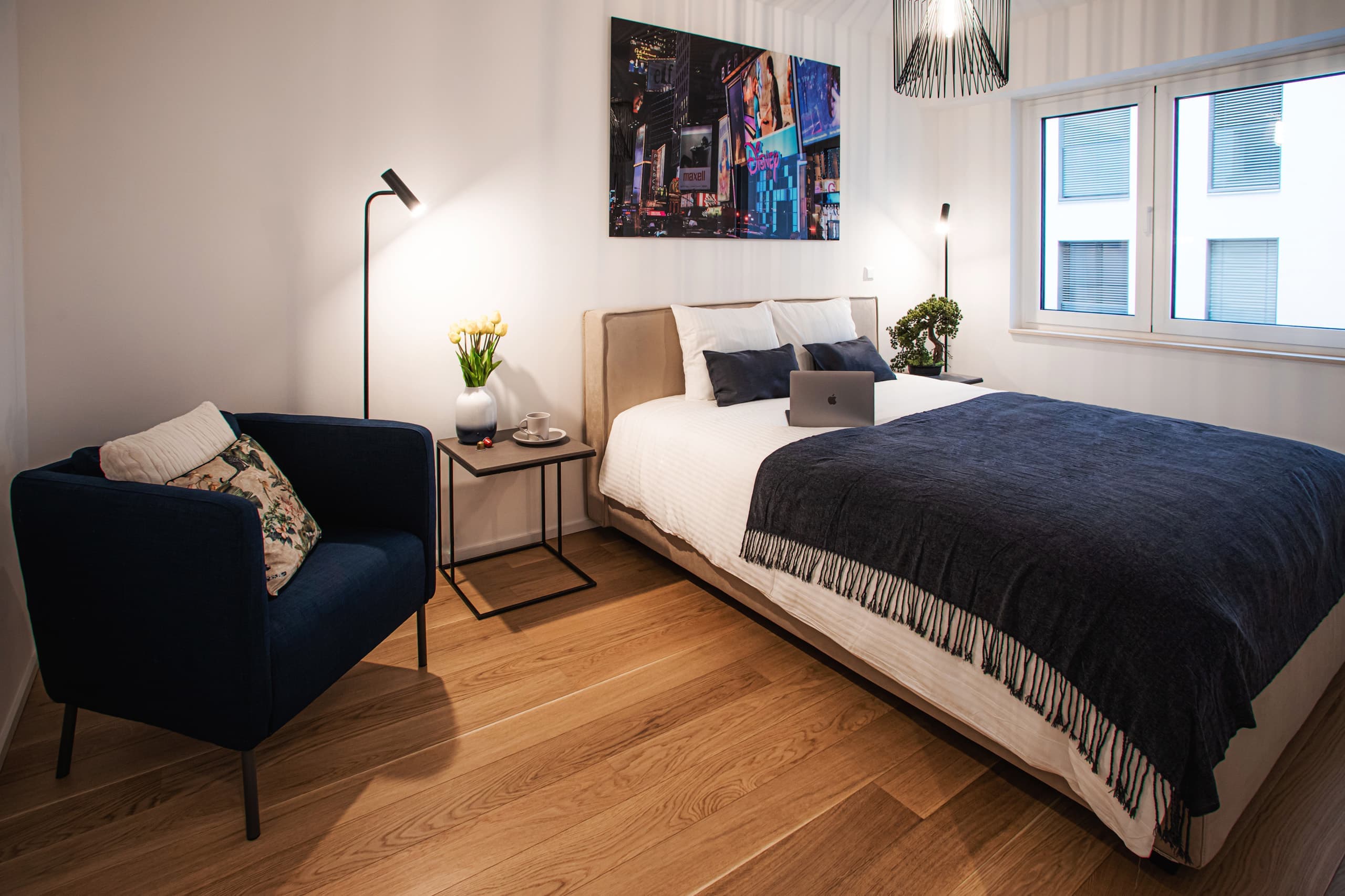 interior of bedroom in apartment Attert at Hamilius Apartments showing a made bed with a macbook pro open sitting on the bed. Next to the bed there is a side table with a vase of yellow tulips and a lounge chair to the left.