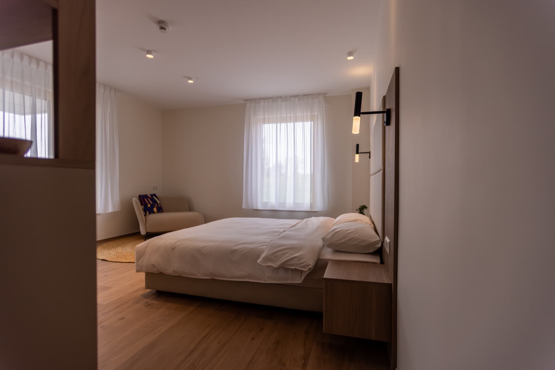 Bedroom with a king-size bed with high-quality white linens. Above bed, there are 2 warm-light lamps.
On the right, there is a beige armchair and large windows
