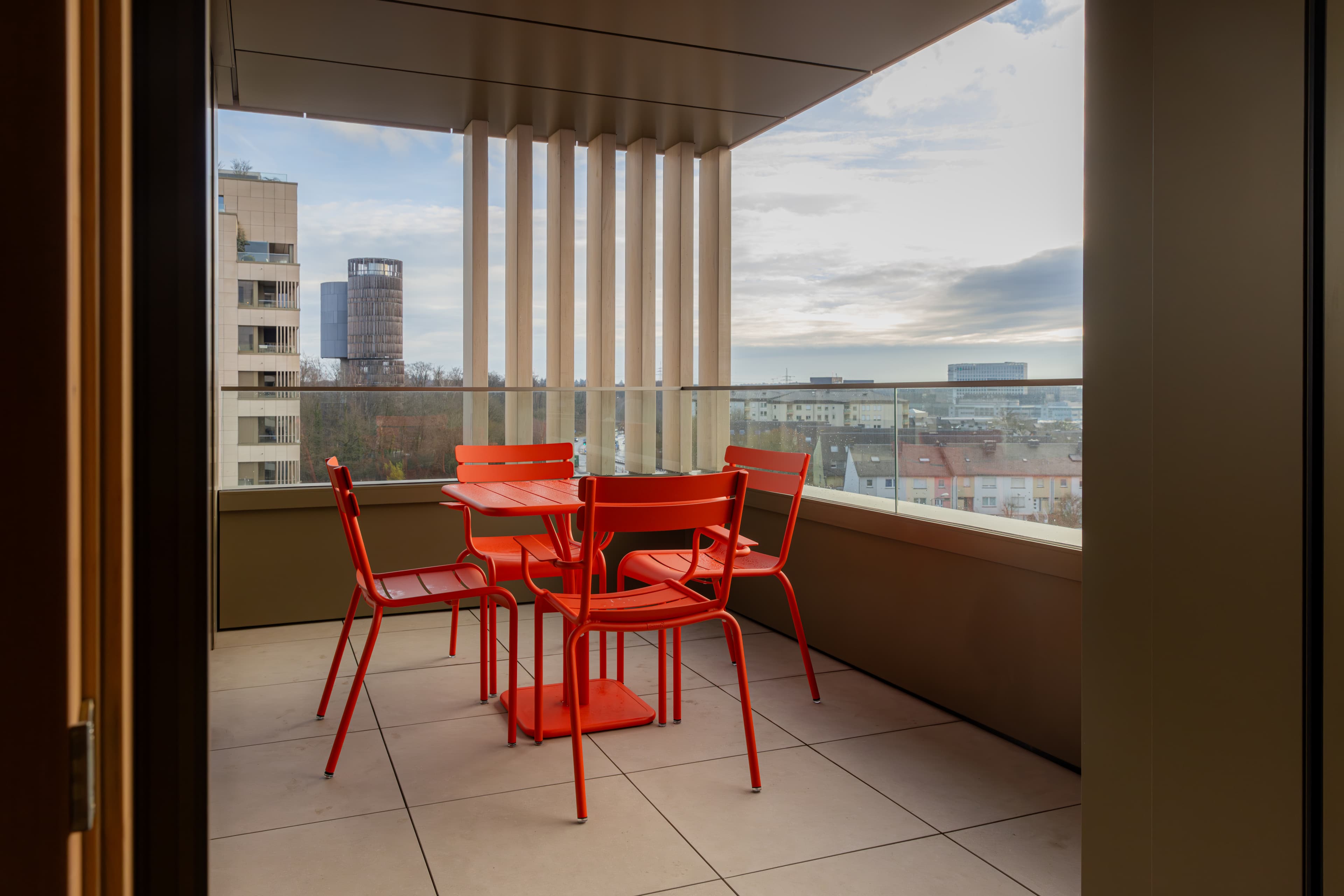 Balcony view of the two-bedroom apartment. It contains a red table and 4 red chairs. The view overlooks the city