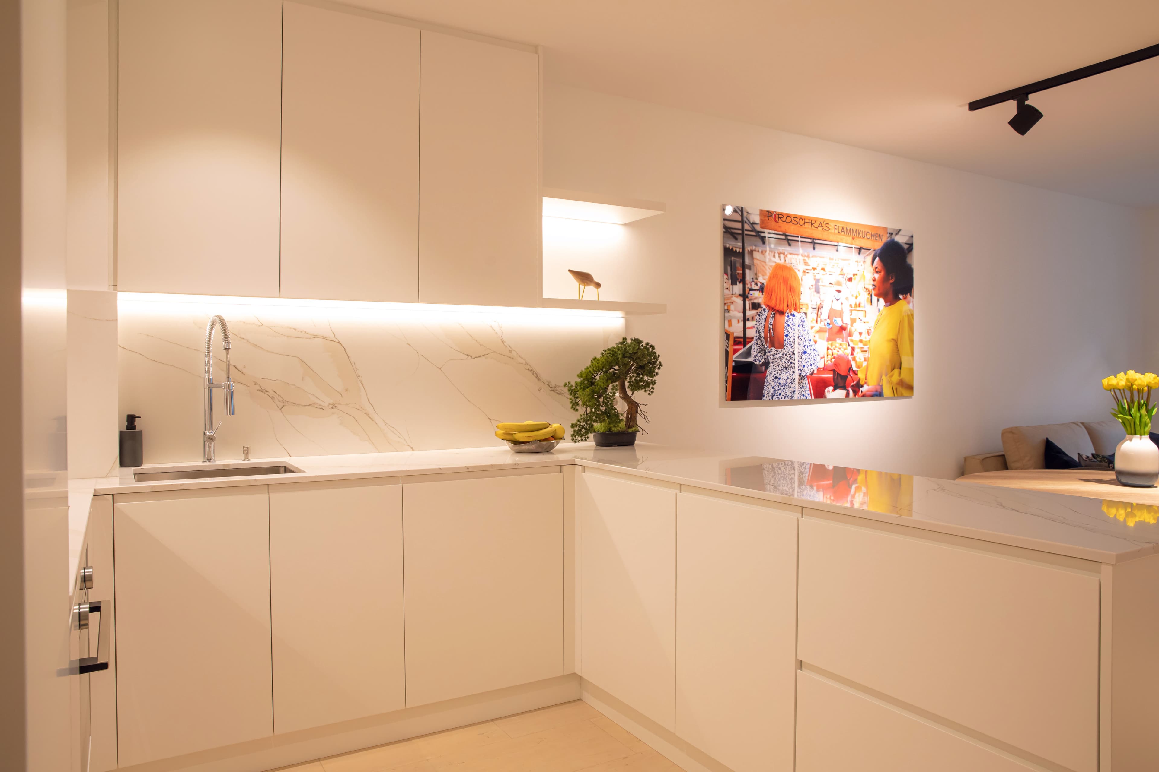 Fully equipped kitchen with a fruit bowl and small bonsai on the counter. In the back, hangs on the wall a big picture of two women in a marketplace.