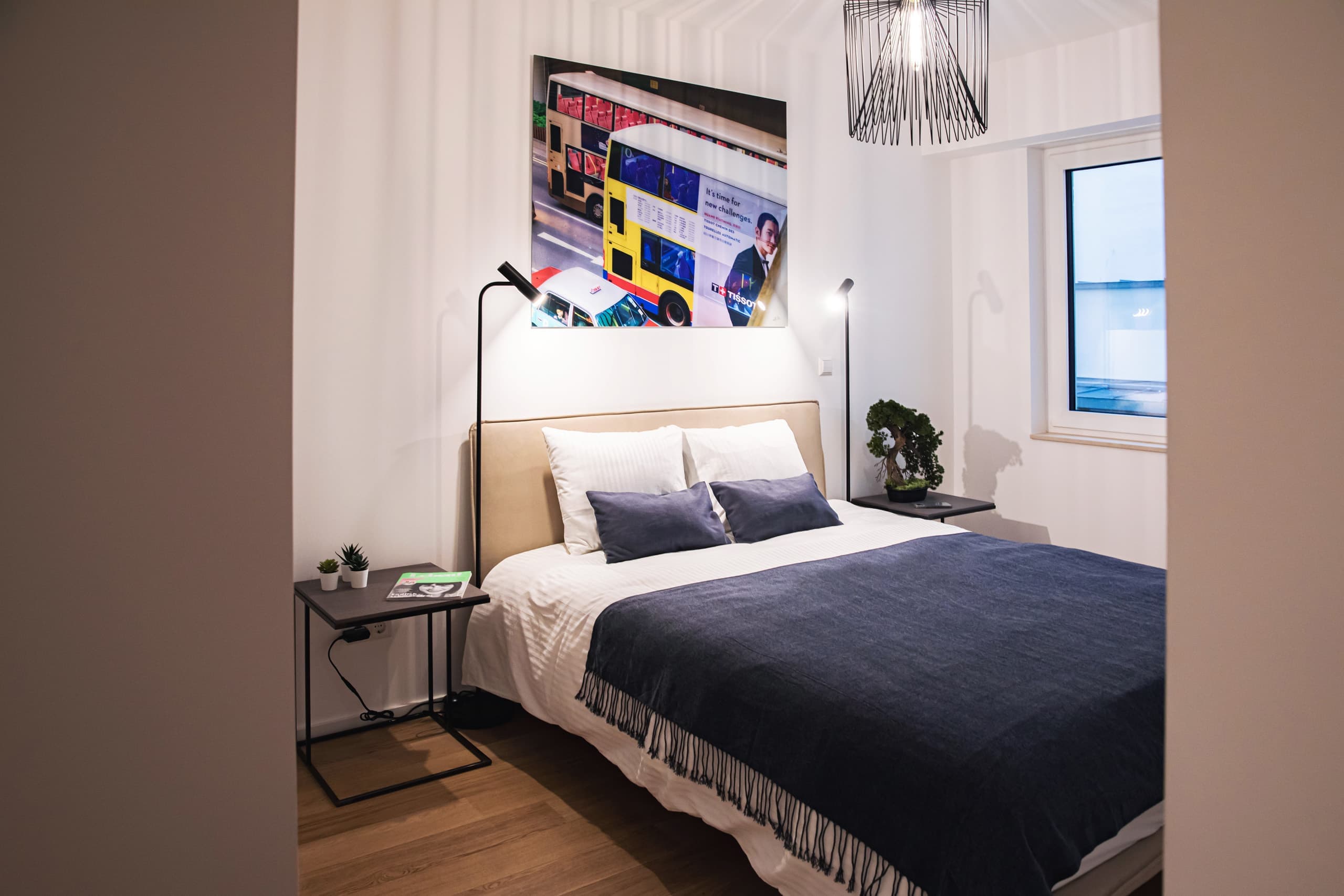 Bed with white and blue sheets and cushions, bedside tables with lamps. Above, is a busy city picture, and on the right, a window.