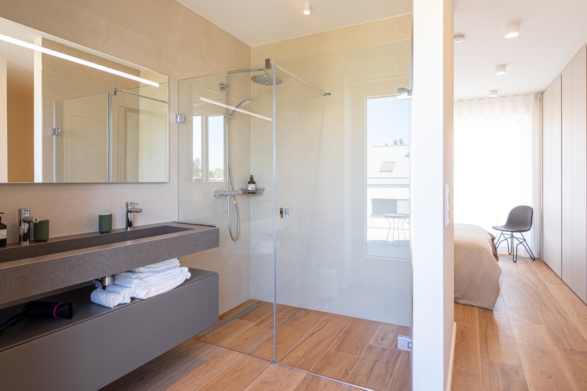 Private bathroom with shower-cabin and sink. Above the sink is a mirror and bellow towels. All amenities included. In the background you can see the bed in the main bedroom.