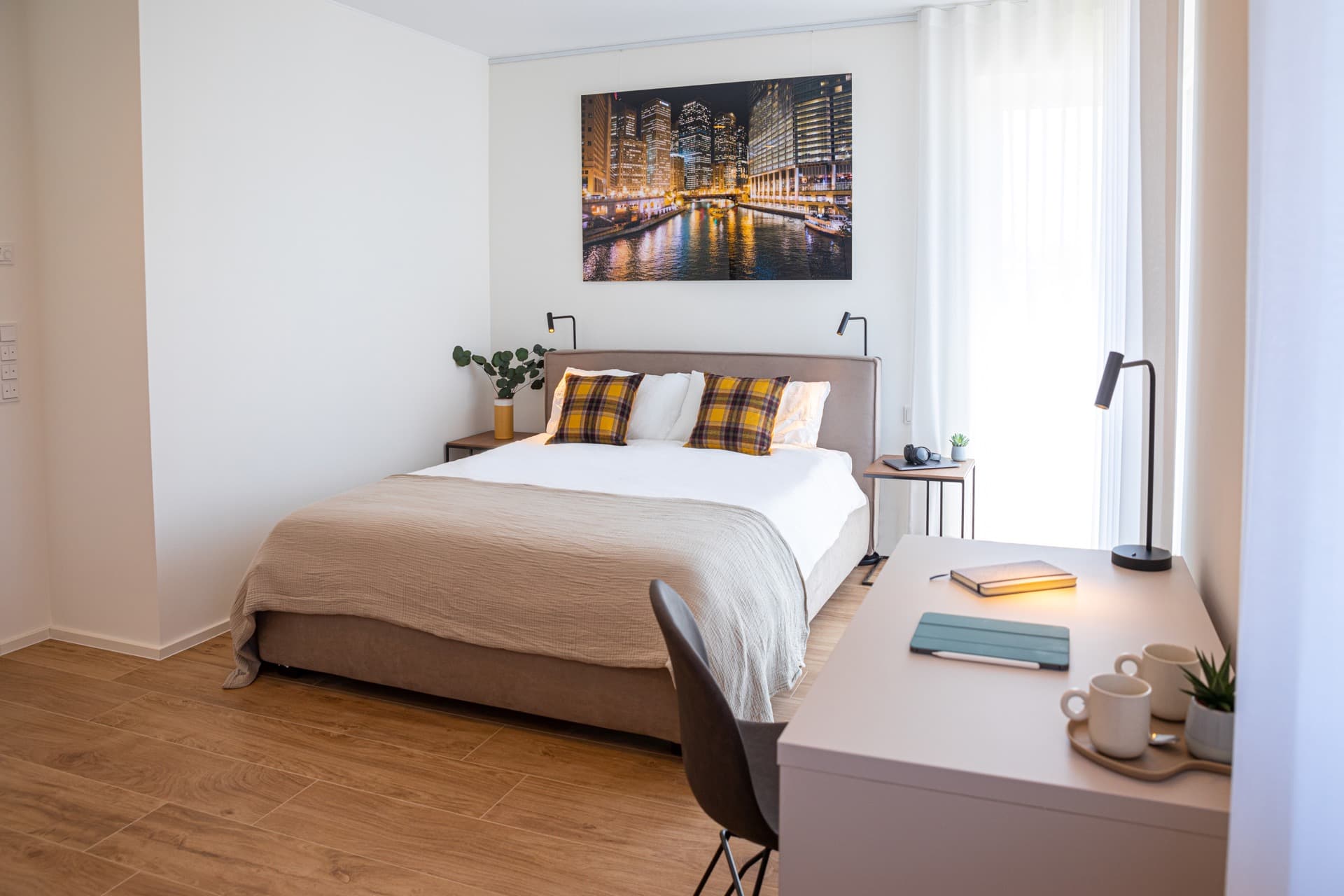 Image of bed with white sheets, cushions, flanked by lamps and nightstands. Above the bed, hangs a picture of a marina. Full-length window with white curtains on one side, desk with lamp, tablet, and agenda on the right.