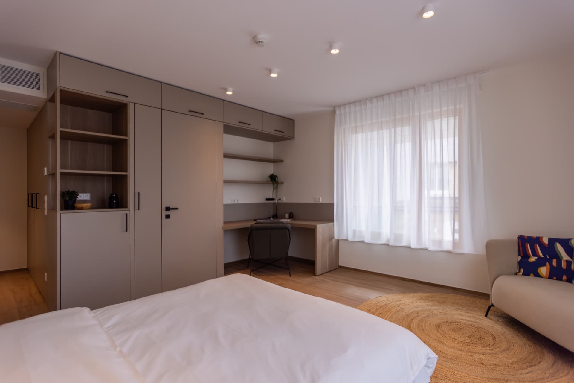 Overview over the bedroom which includes: a king-size bed, a designated working area, storage room and dressing, and large windows flooding the room with natural light.