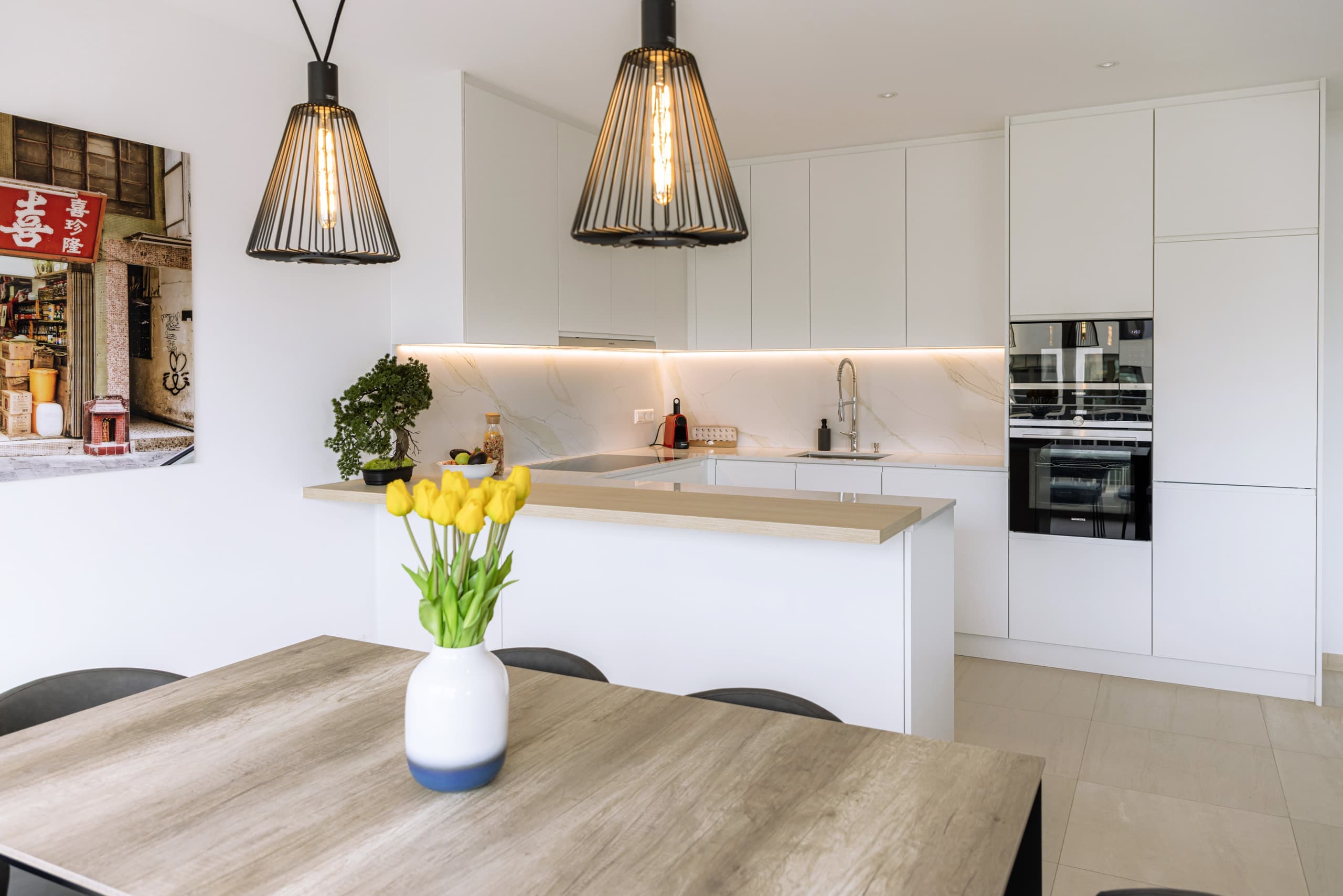 In the foreground a round dining table with chairs, and with a flower vase with yellow tulips. In the background, a fully equipped kitchen with a coffee machine and a bonsai flower pot.