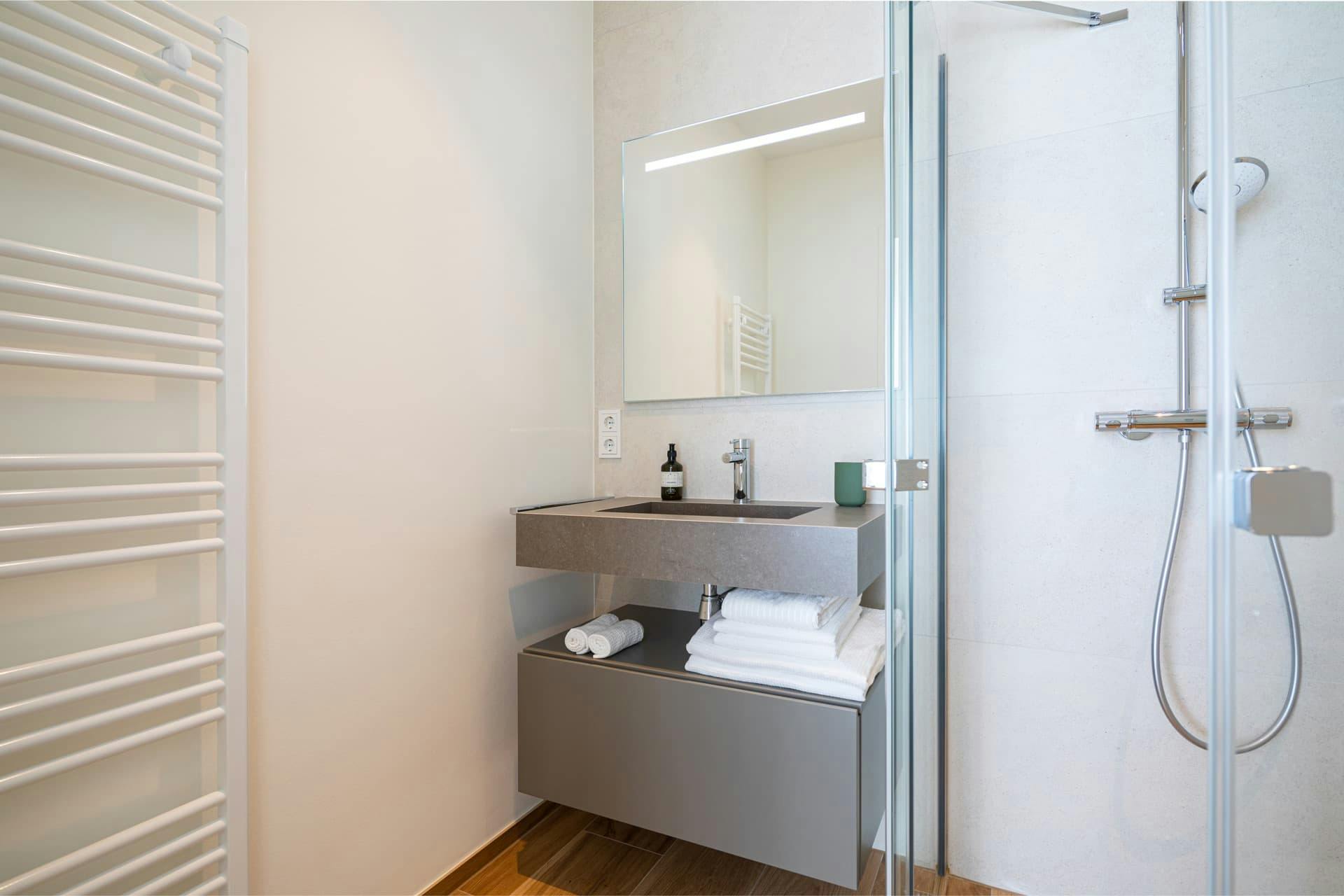 Modern sink with mirror above, white towels below. Part of the shower is visible on the right. Two electric sockets are on the left. Reflected in the mirror is a heating radiator. 
