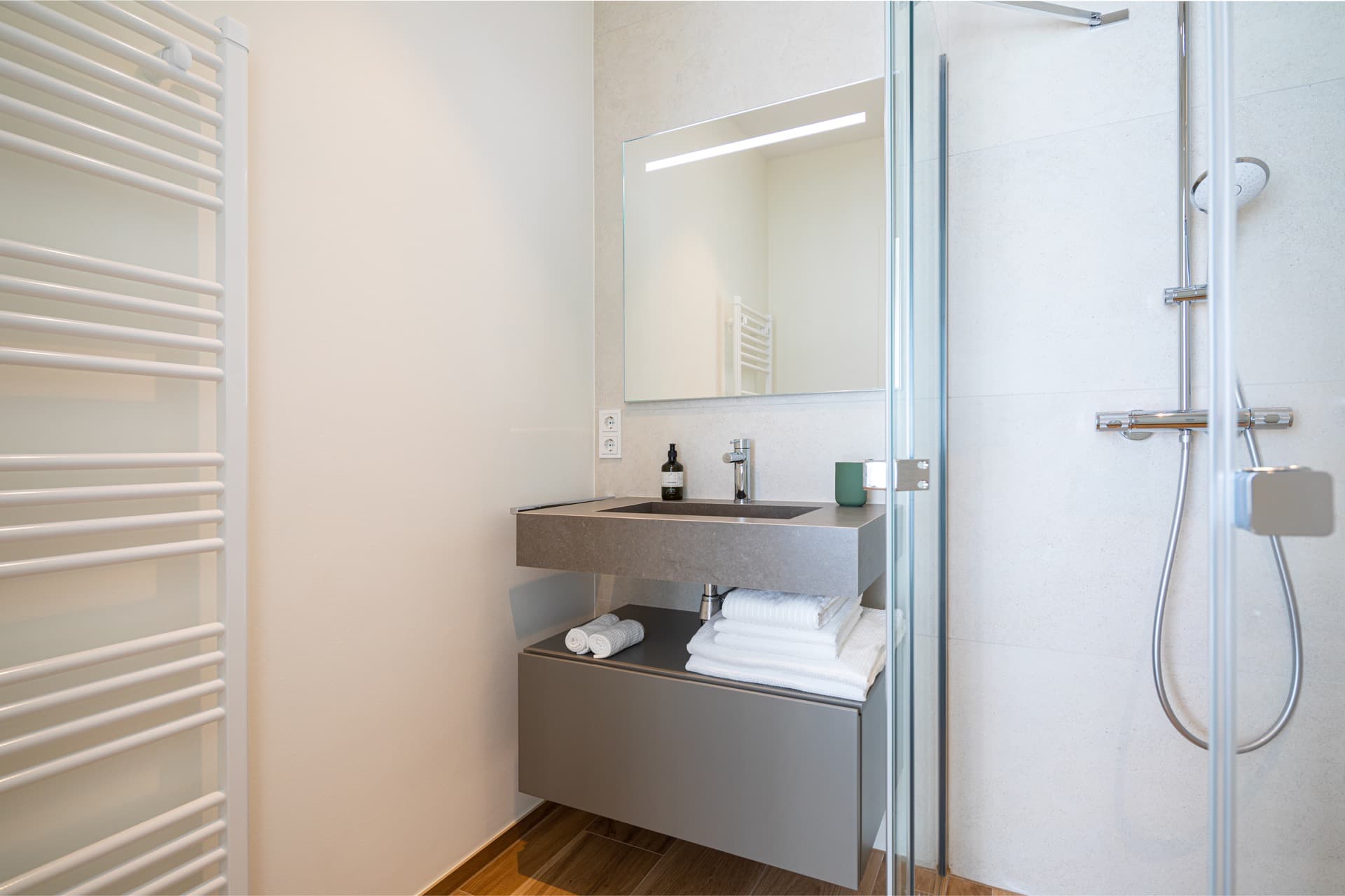 Modern sink with mirror above, white towels below. Part of the shower is visible on the right. Two electric sockets are on the left. Reflected in the mirror is a heating radiator.