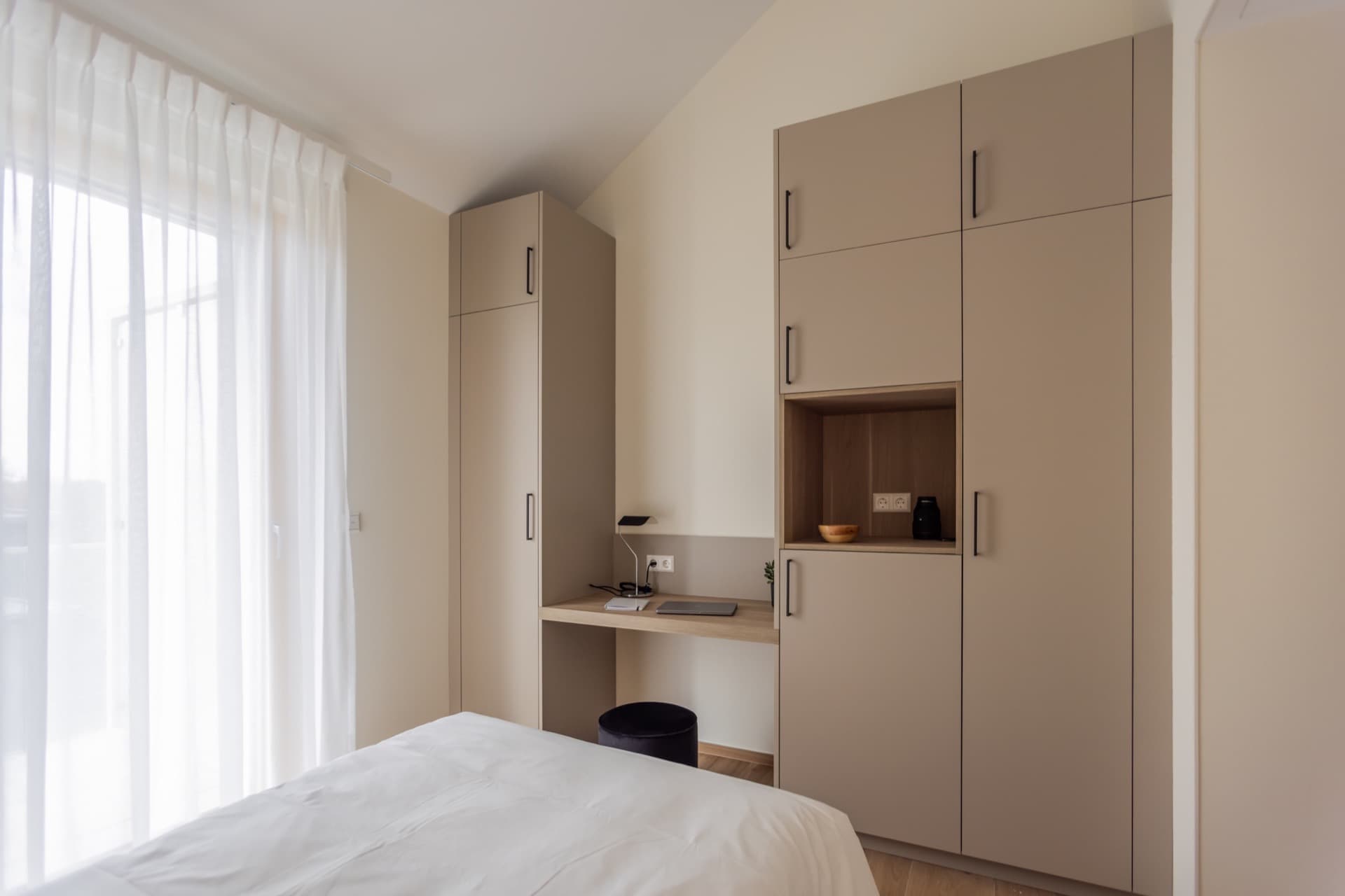 The picture depicts the bedroom's working area. It has a desk, storage rooms, and drawers attached. On the desk there is a laptop and a lamp.
On its left, there is a large floor-to-ceiling window with curtains.