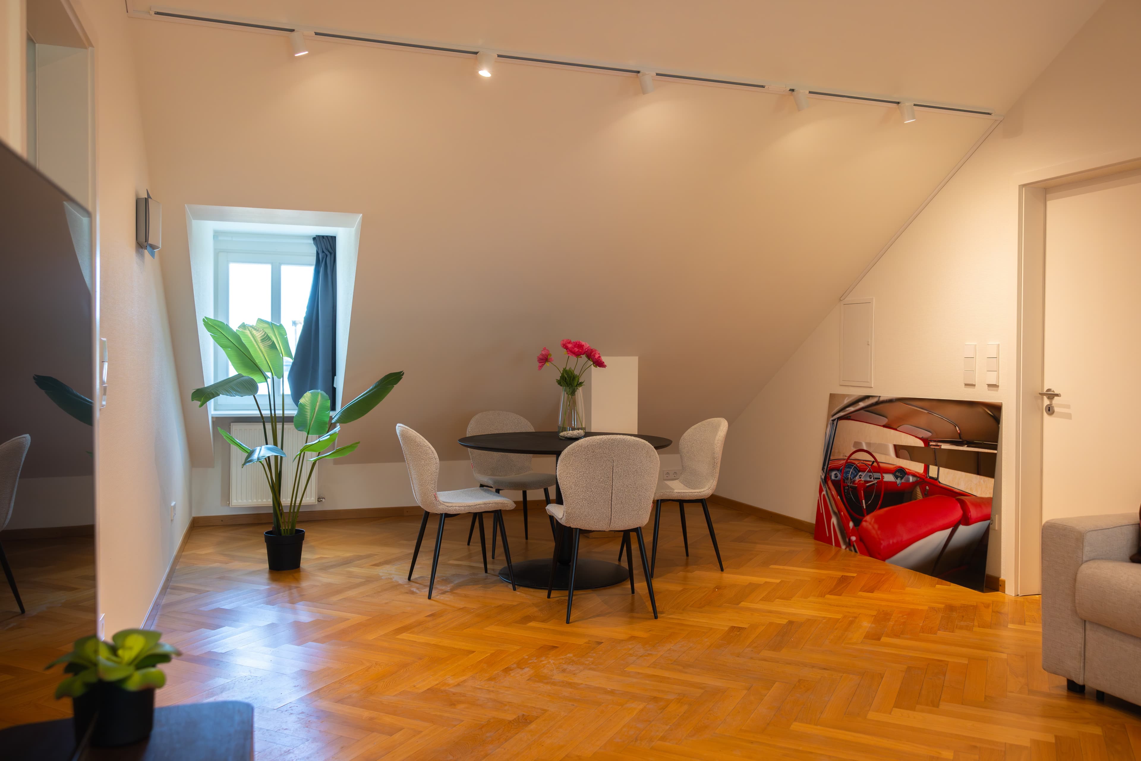 The picture depicts a table with four chairs, a wall grand picture, and a flower pot.
In the background, there is a window with a curtain, and a heating radiator underneath.