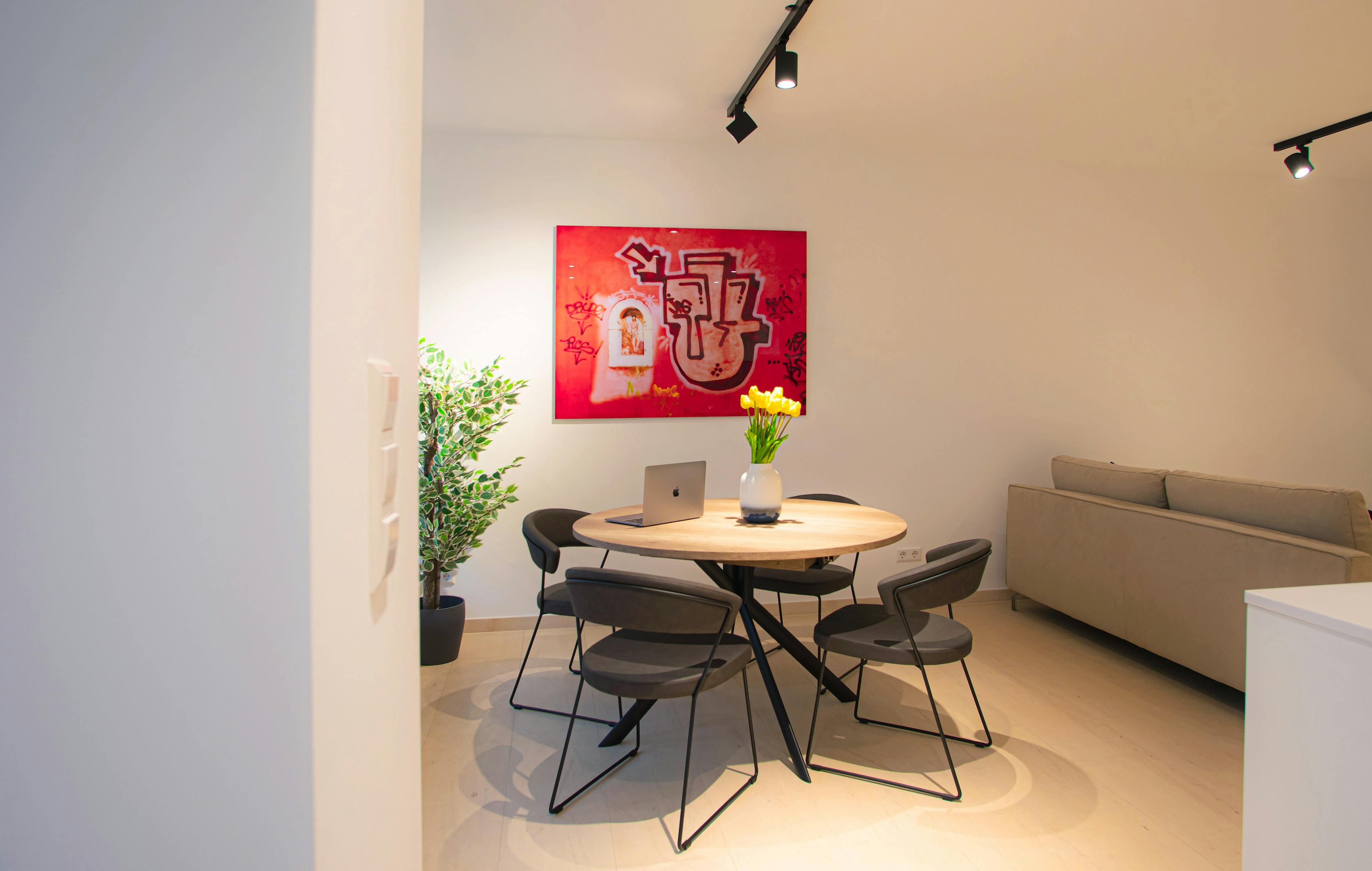 Round dining table with chairs, next to the sofa, on the right. On the left, there are big windows and a big flower in a pot. In front of the table, there is a big red picture hanging on the wall.