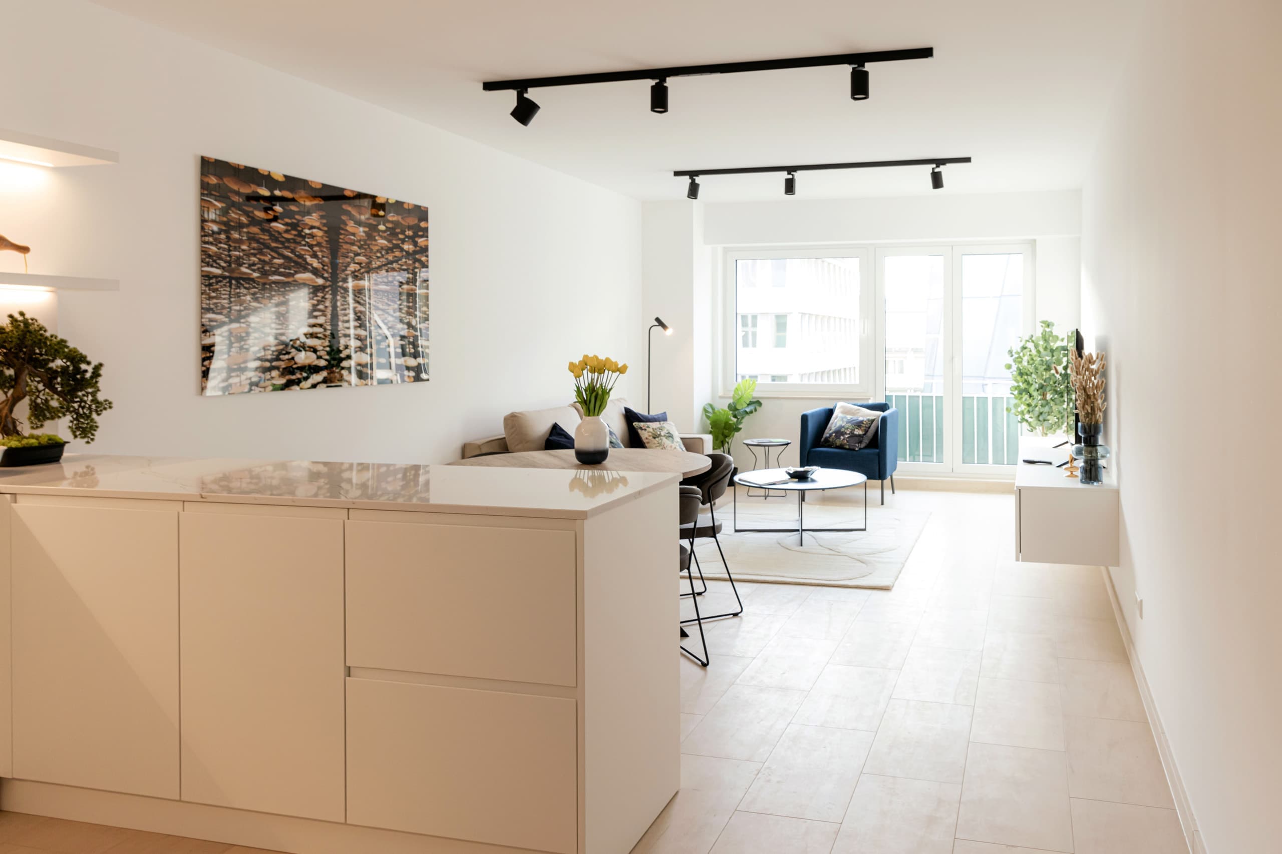 Overview over the kitchen countertop and the lounge area. In front of the kitchen, there is a round dining table with chairs and a flower vase on top. Next, there is a couch with cushions, and, in front of it, on a white carpet, there is a medium-sized coffee table. In front of the large windows there is an armchair.