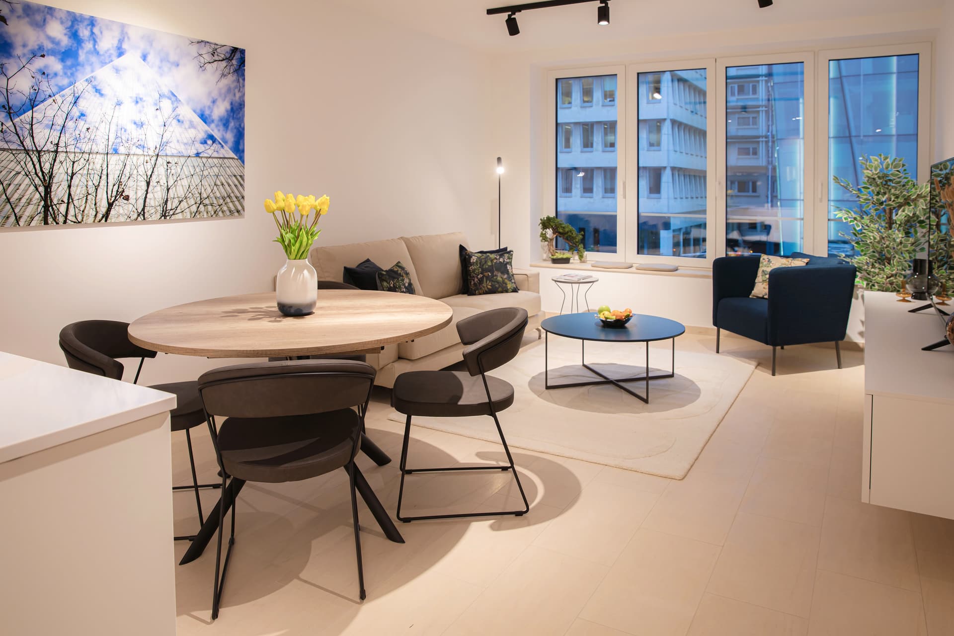 The image shows a circular living room table decorated with a vase of yellow tulips and three chairs. To its left is a couch with cushions, and in front of it, a small coffee table with a fruit bowl. Positioned in front of the small table is a dark blue armchair. Adjacent to it is a white TV stand with a TV.