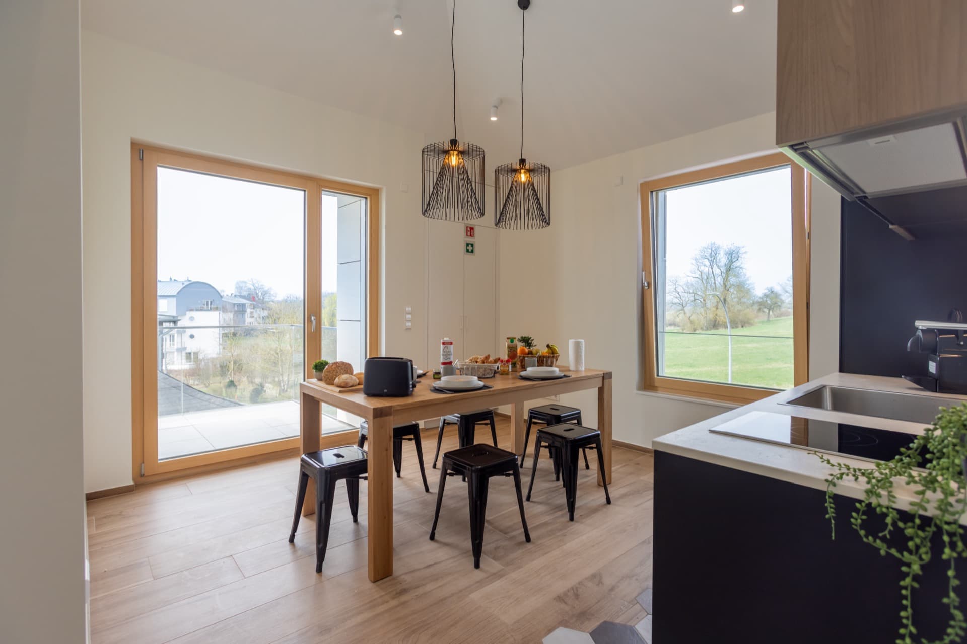 Overview of the shared kitchen space in Cessange House offering rooms to rent in Luxembourg City. There is a tall kitchen table & chairs, large floor-to-ceiling windows and kitchen appliances.