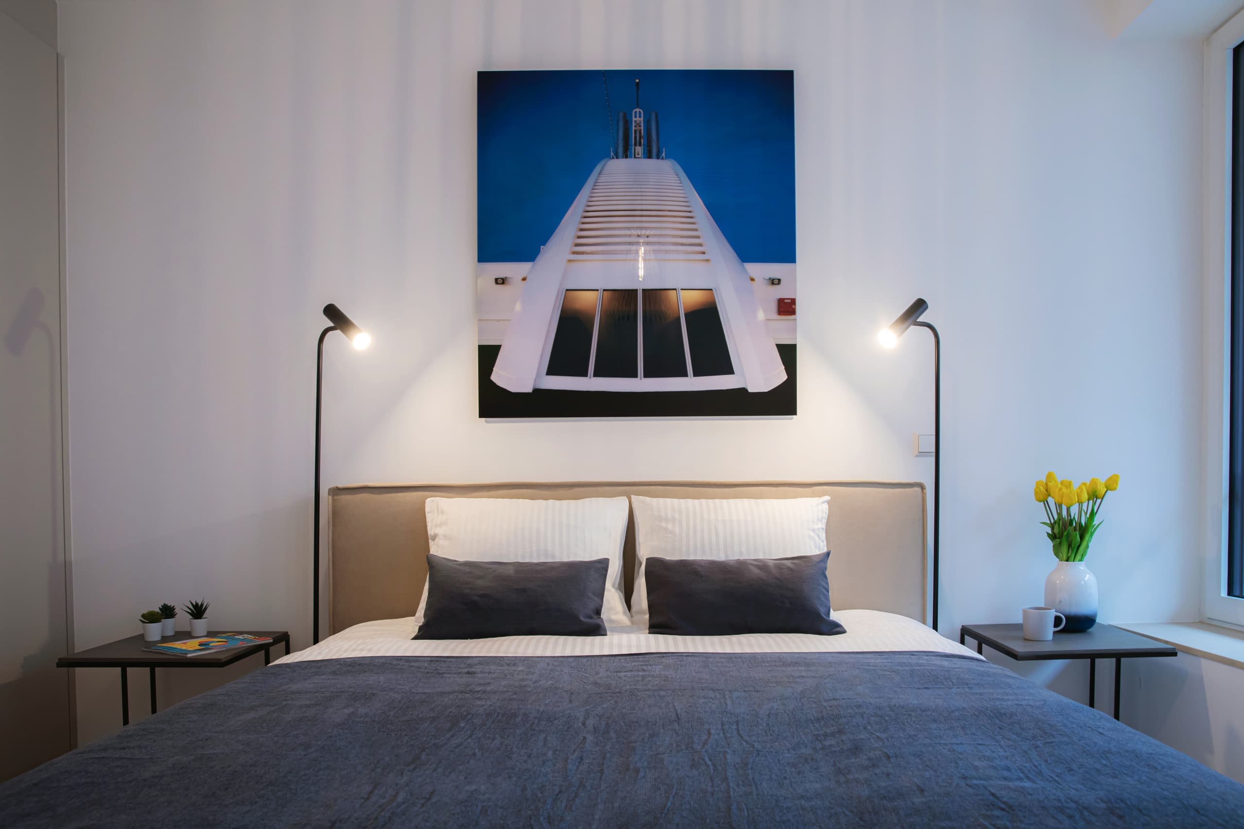 Bedroom with large bed flanked by two lamps and two nightstands. Behind the bed, on the wall, hangs a big picture. On the right, there are windows.