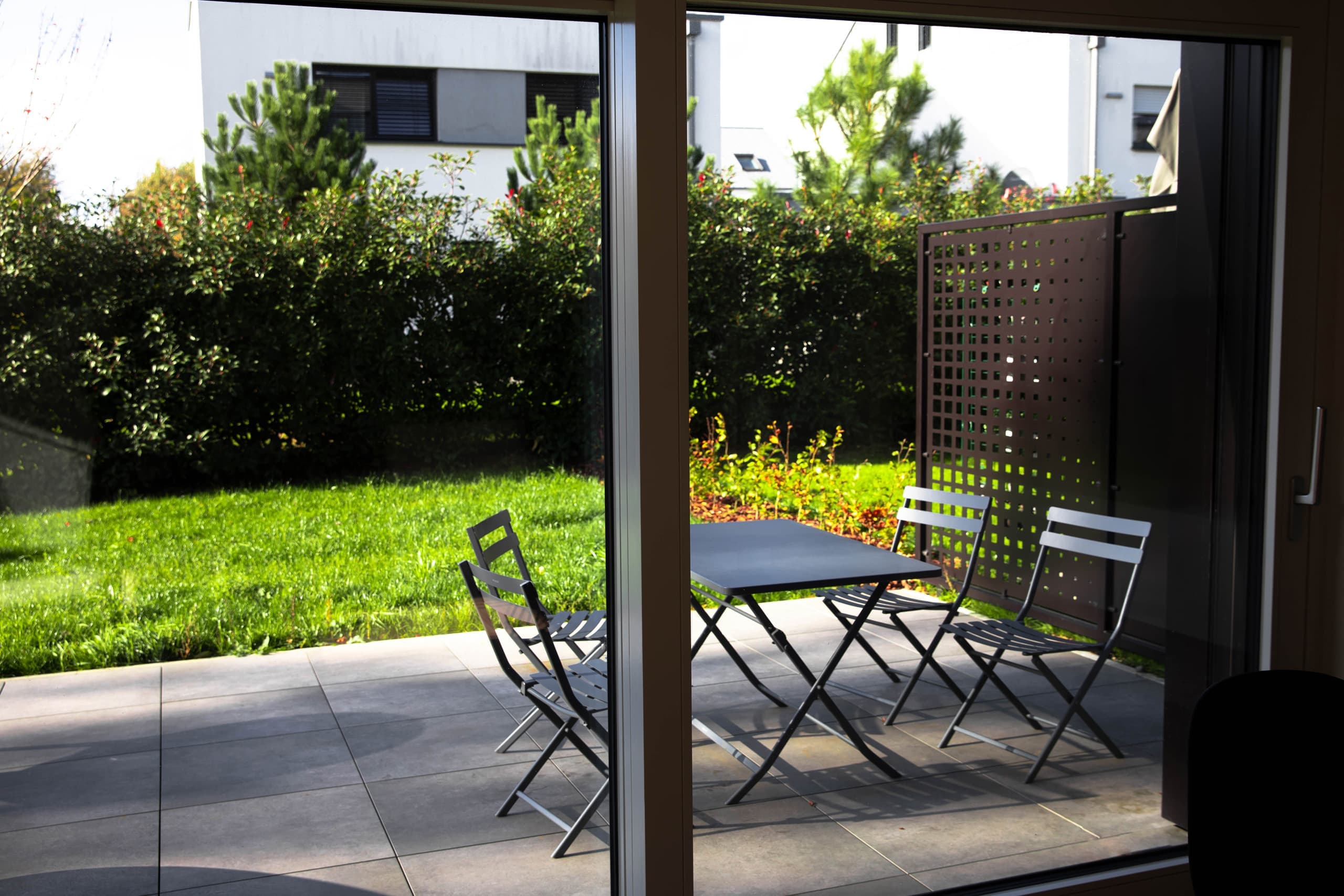 The picture illustrates the view from the inside of the house, showing the exterior dining area on the terrace with a table and chairs, and the garden behind.