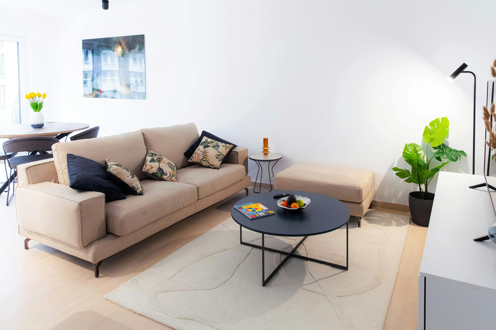Sofa with cushions on it. In front, is a medium-sized coffee table, a TV stand with a smart TV on it, and next, a small armchair. Behind the sofa is a dining table with chairs, next to large windows. On the wall, hangs a big picture.