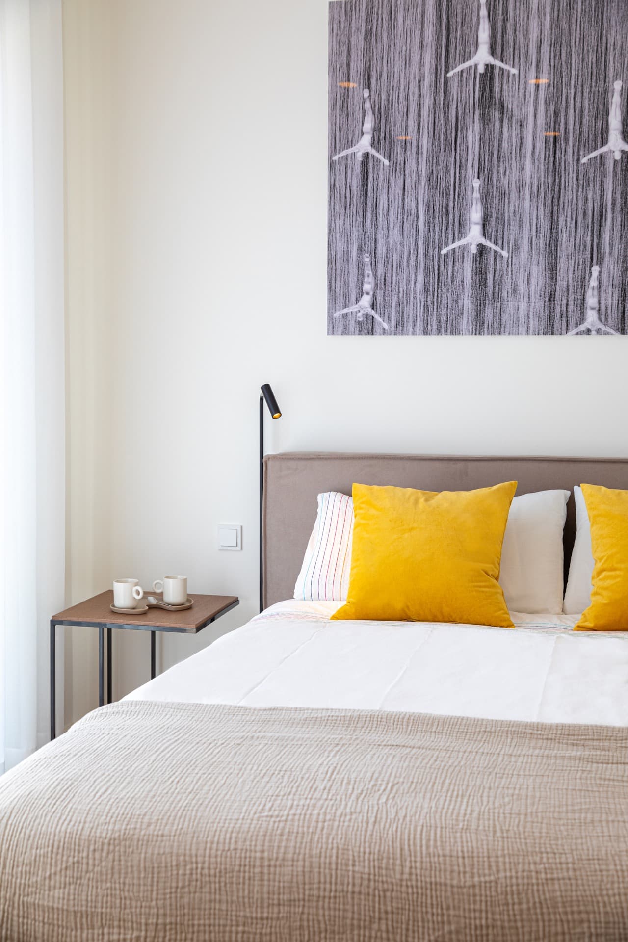 Close-up image of the bed with white sheets and mustard pillows. On the left, a standing lamp and a nightstand. Light coming through the big windows.