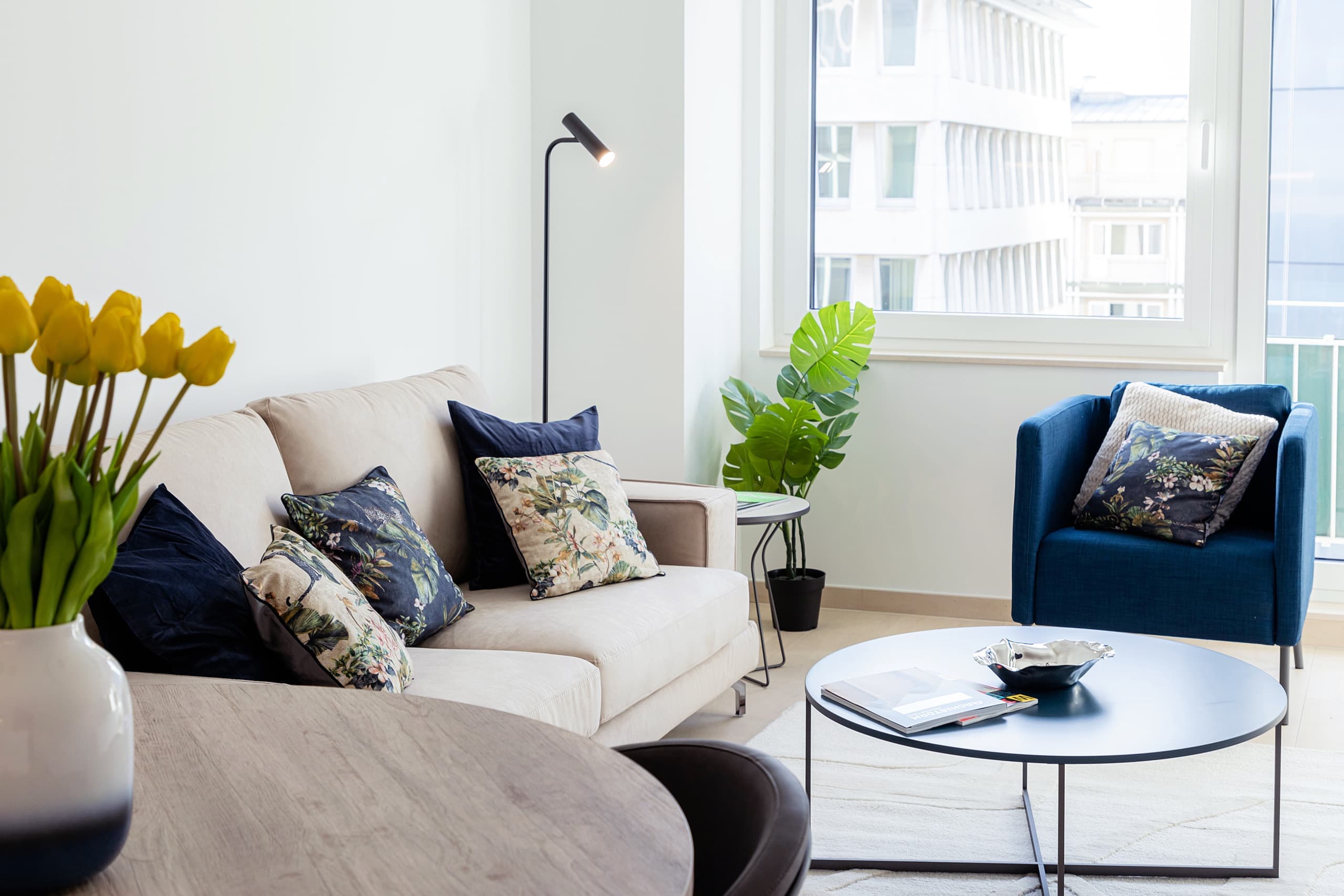 Close-up image of the lounge sofa, with multi-colored cushions on it. In front, there is a medium-sized coffee table, and next to it, there is a dark blue comfortable armchair. A big flower pot can be seen under the windows.