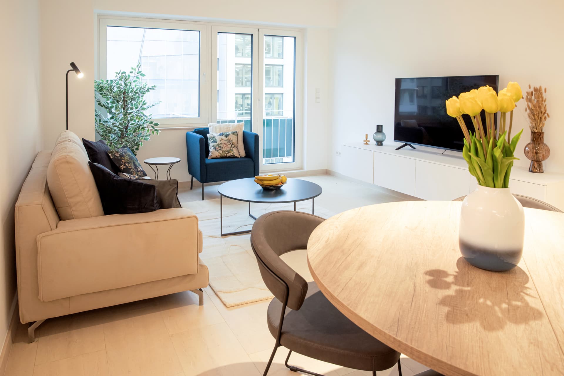 Round dining table with chairs and flower vase on it. On the left, there is a couch with cushions, and, on the right a white TV stand with a smart TV on it. In the center of the room, there is a coffee table and an armchair. In the background, you can see the balcony door and the view.