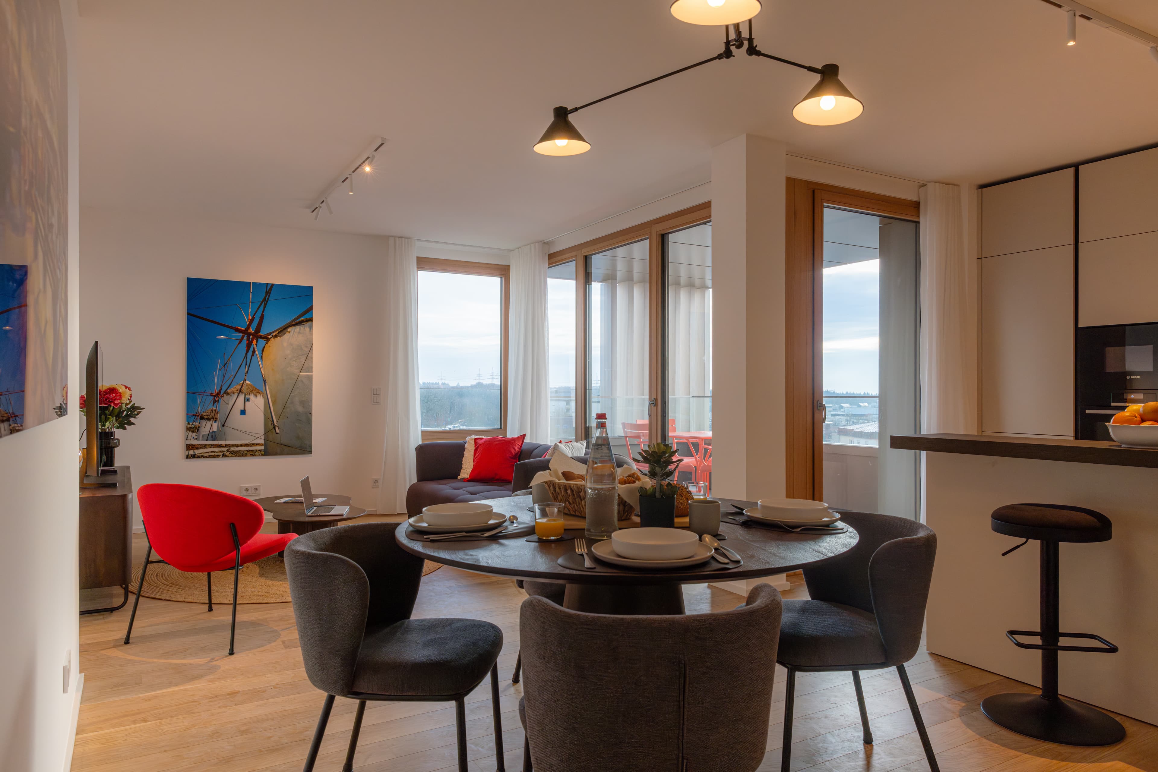 Overview of the lounge and dining area of the apartment.
There is a dining table with chairs, a sofa, a coffee table, a red armchair, and a TV stand with an LCD TV. Behind the couch, there is a large terrace with a red table and 4 red chairs.